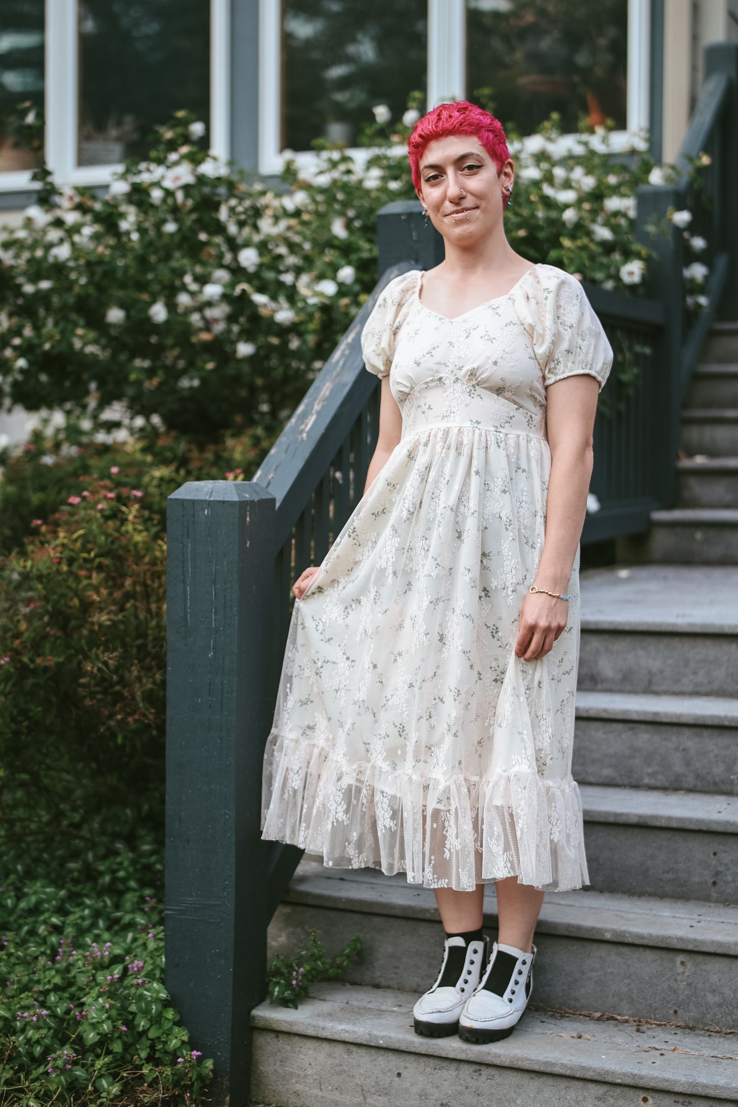 A person in a floral white dress and pink hair stands on outdoor steps wearing casual sneakers.