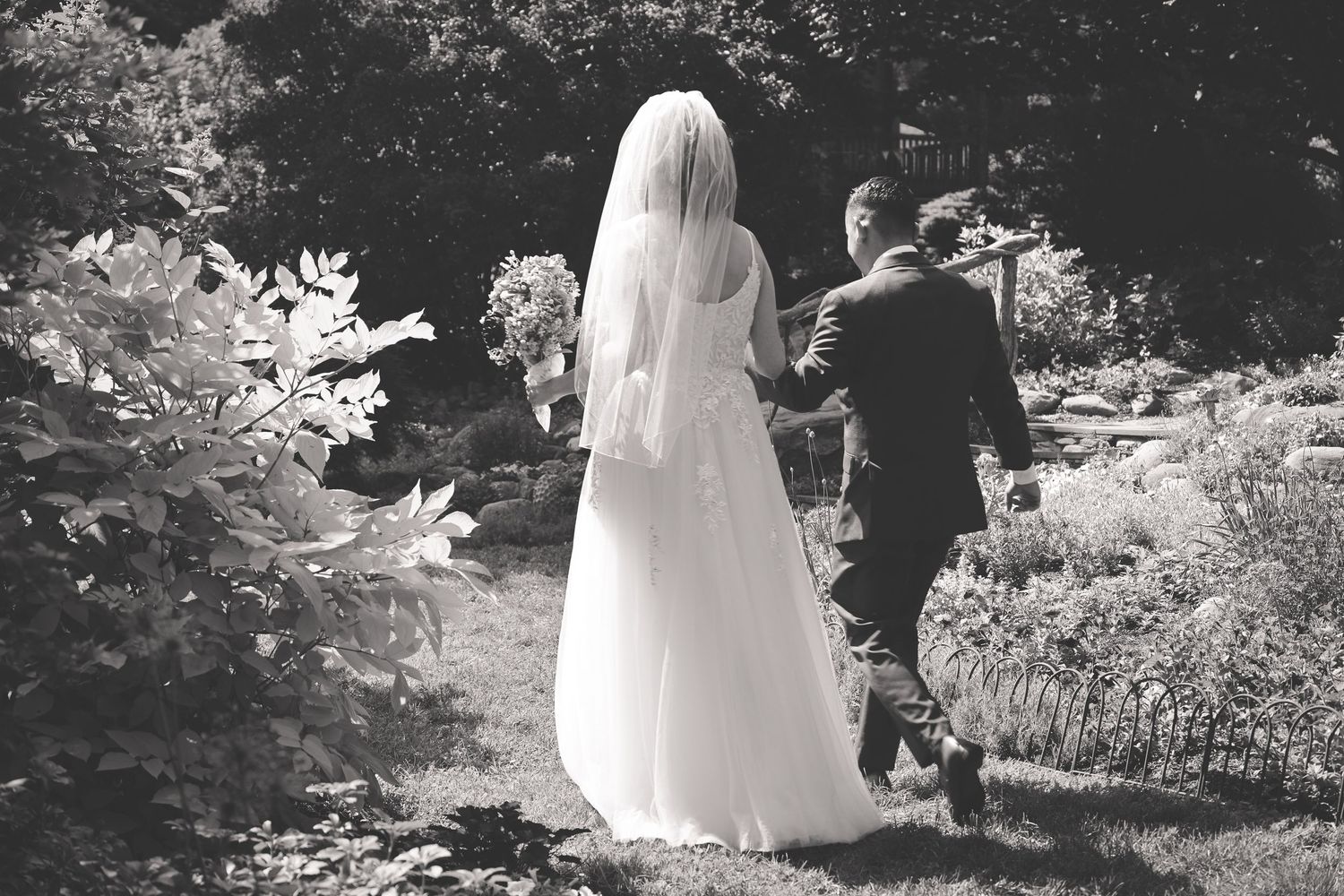 A couple in wedding attire walks hand in hand through a lush garden path on their special day.