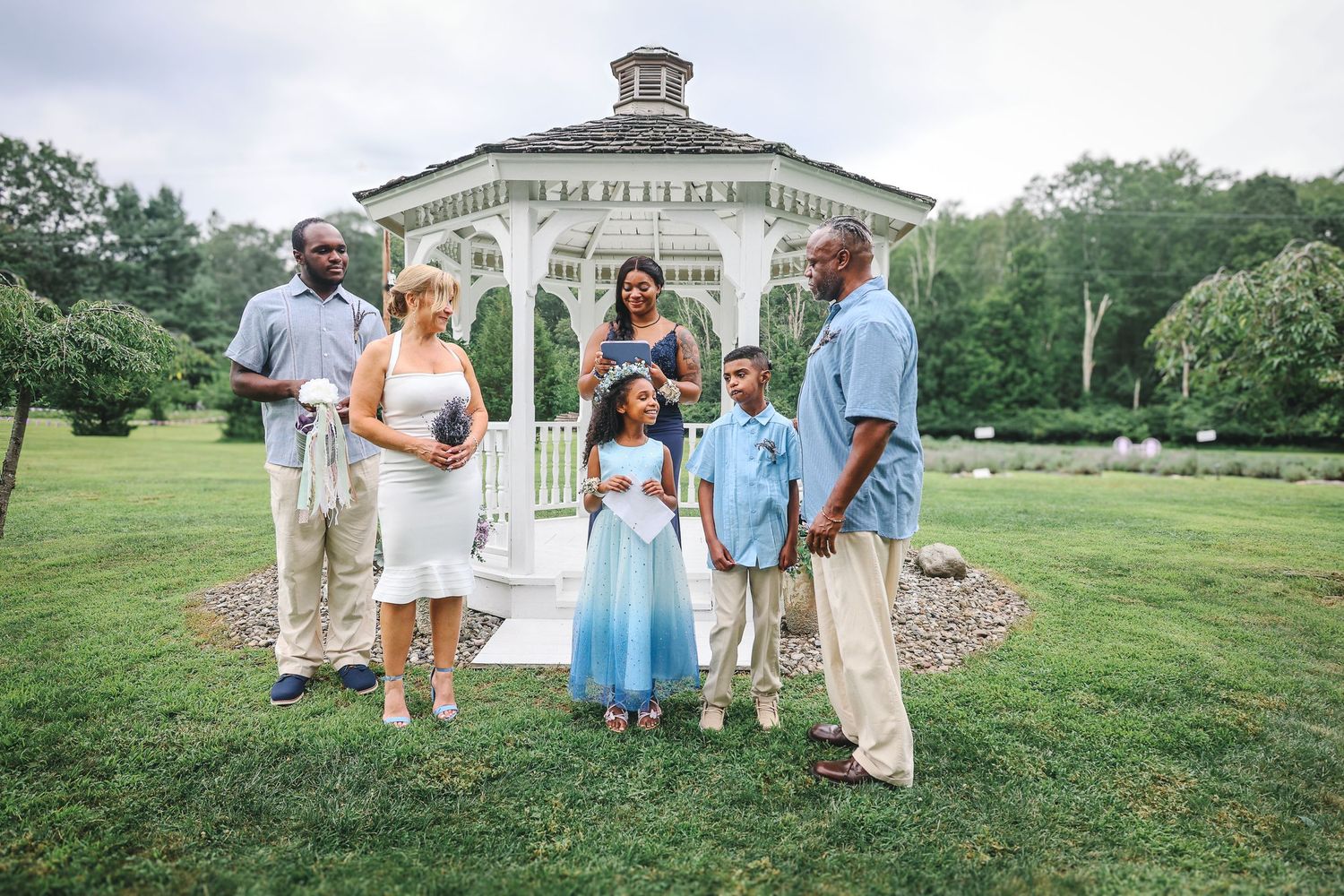 Wedding party in light blue and white attire poses together on a grassy hillside during an outdoor celebration.