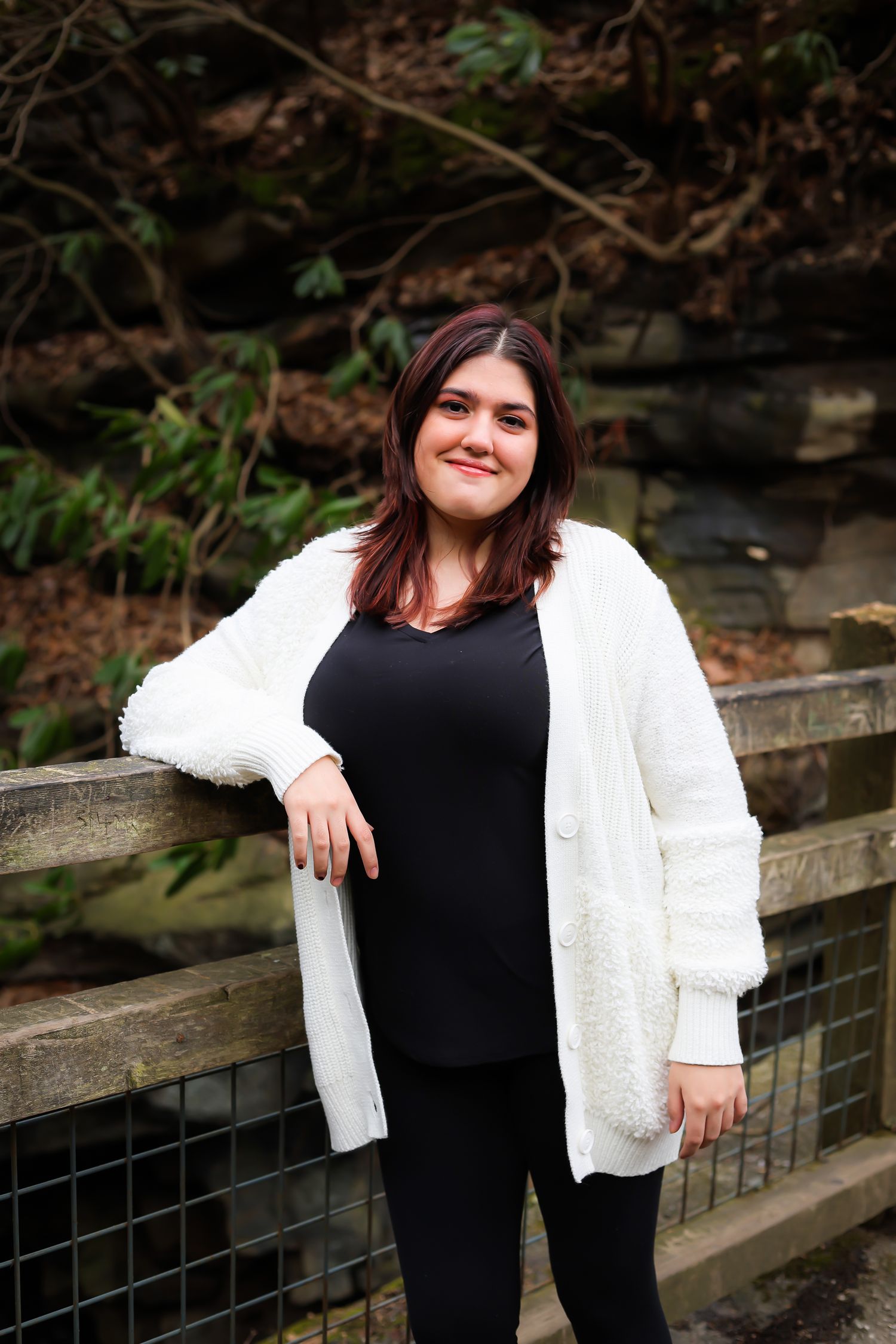 Person in a cozy white cardigan and black outfit poses on wooden steps surrounded by greenery in an outdoor setting.