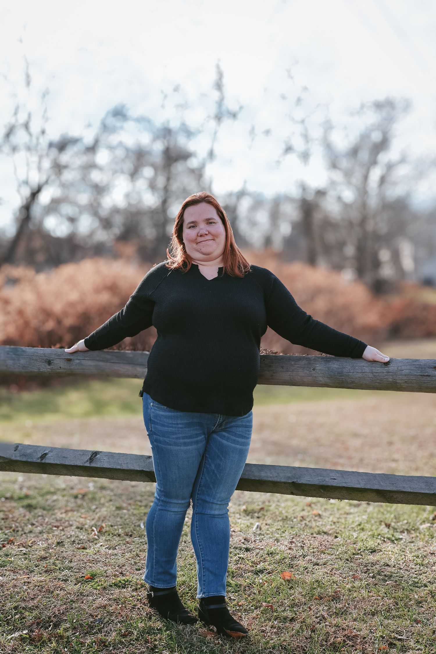 Person in black sweater and jeans stands by rustic wooden fence in autumn field during outdoor photo session.