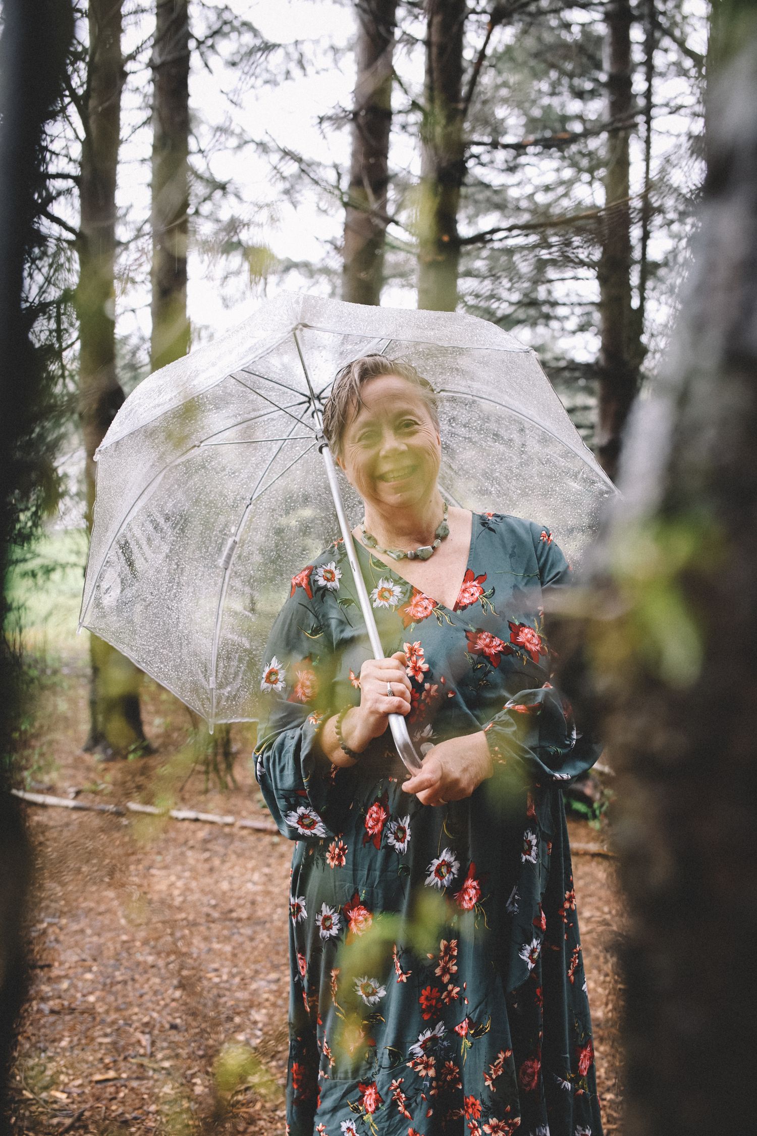 Person in a floral dress holds a clear umbrella while standing among trees in a forest setting.