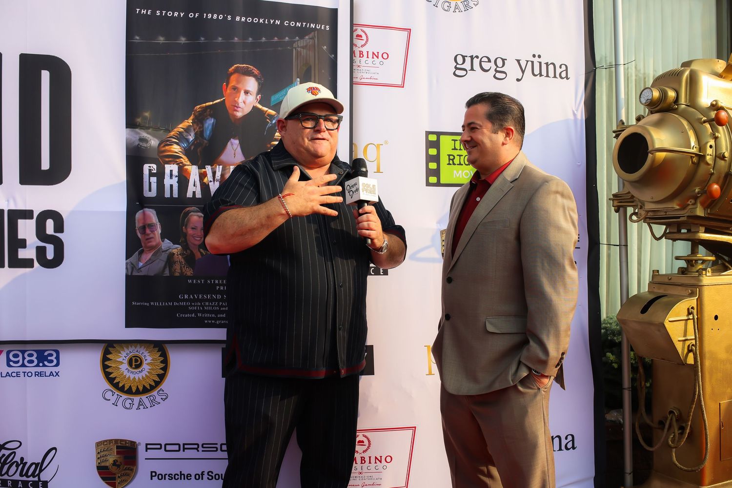 Two people stand in front of movie posters at an entertainment event, with one speaking into a microphone.