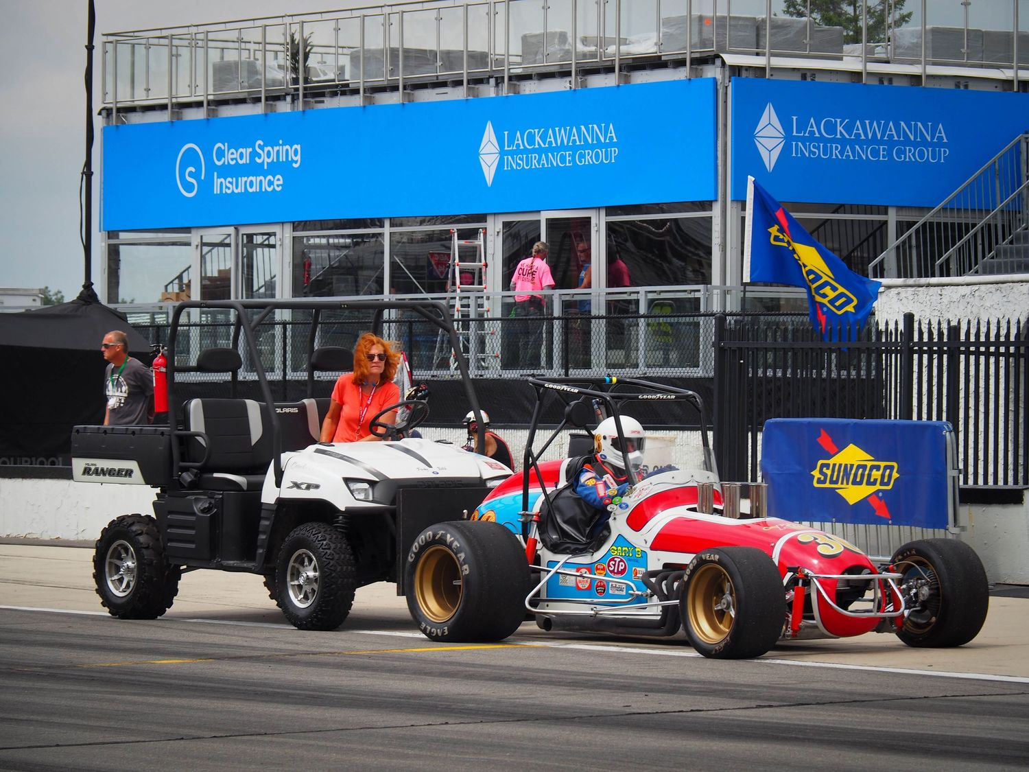 Vintage Formula One race cars line up along a pit lane with blue barriers and signage visible in the background.