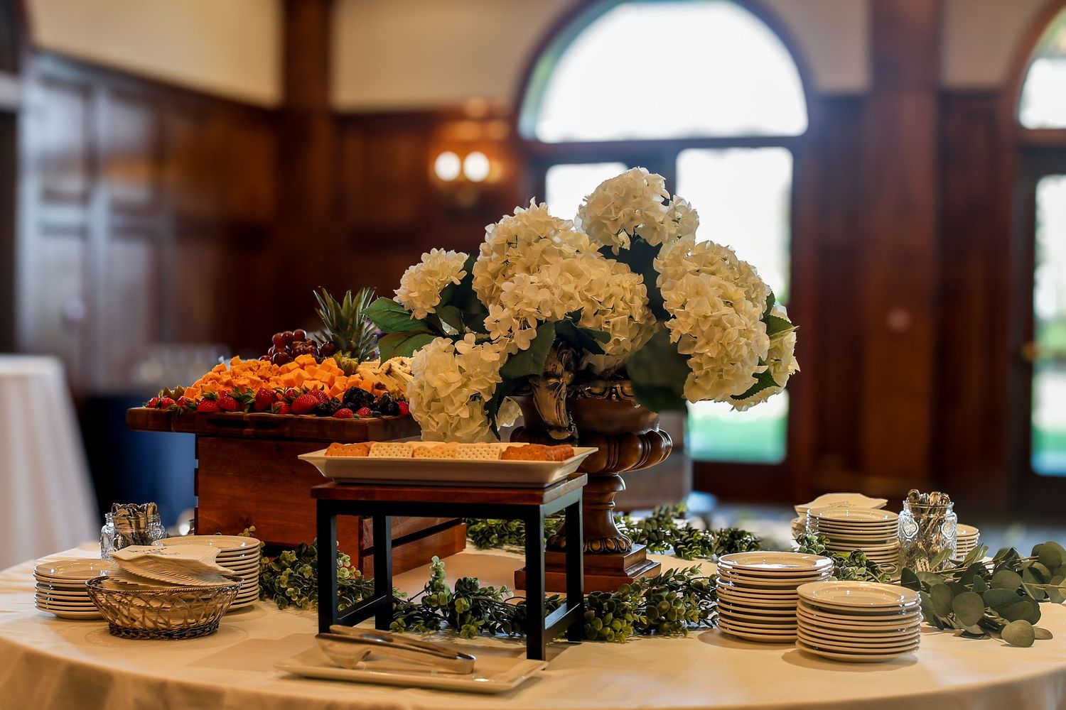 Elegant buffet table with white dishes, floral centerpieces, and decorative food displays in a warm-lit dining room.
