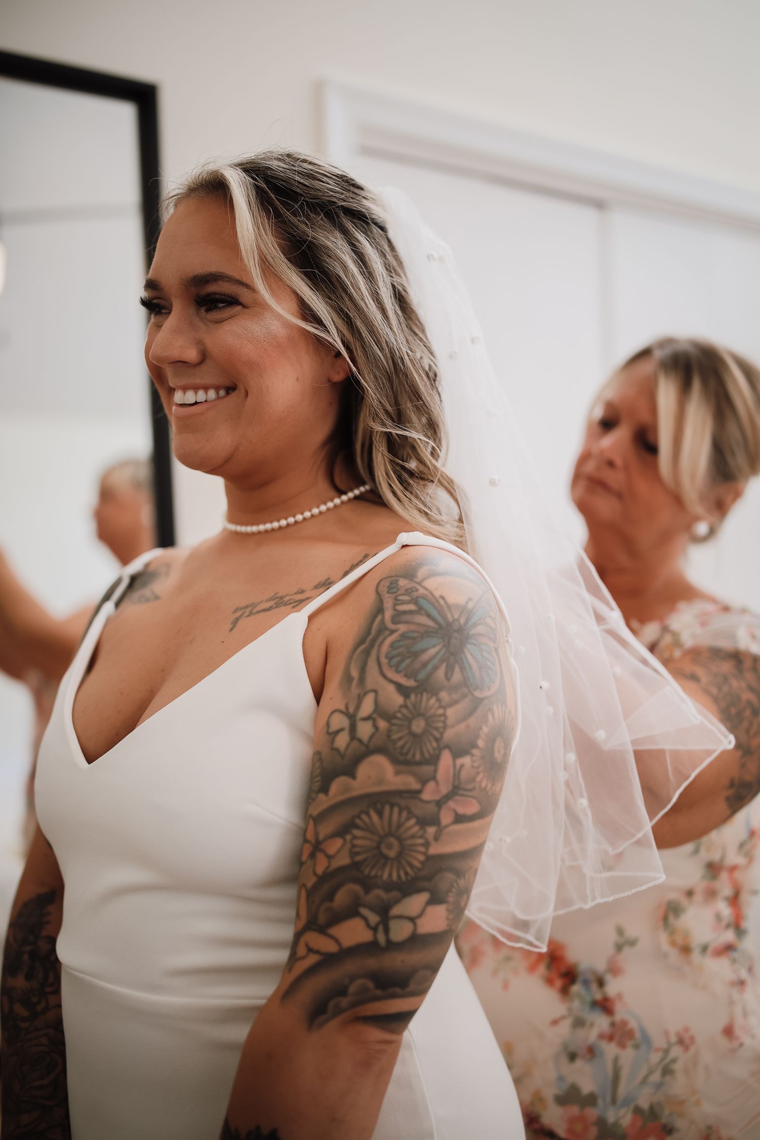 A close-up view of a bride in a white dress with visible shoulder tattoos and pearl necklace during wedding preparations.