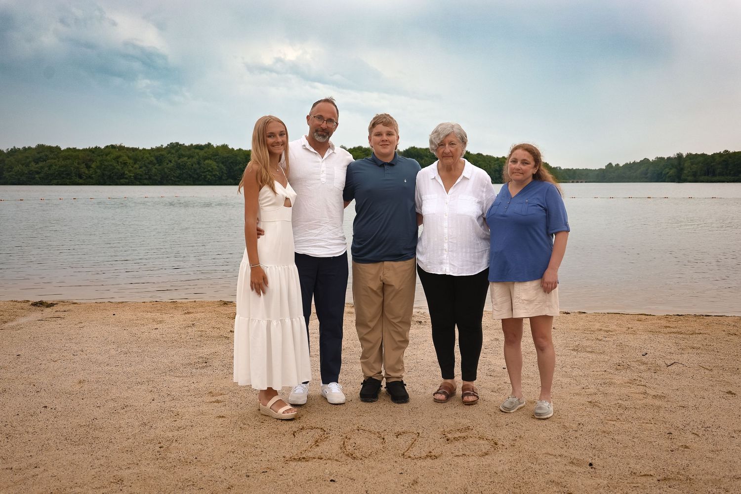 A group gathers for photos on a sandy beach with a lake or ocean view in the background.