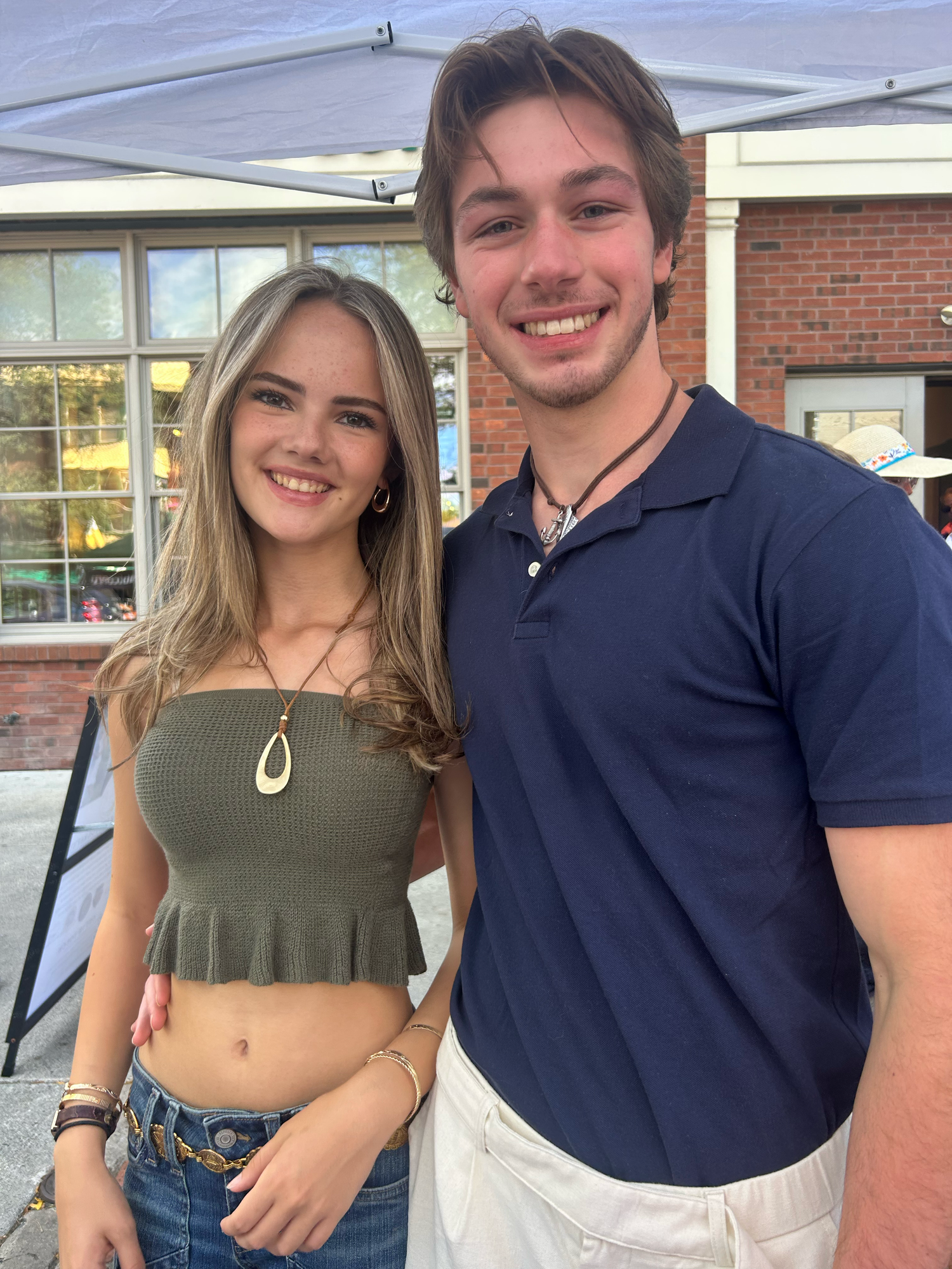 A couple poses together outdoors in casual attire, the woman wearing a crop top and jeans while man in navy polo shirt.