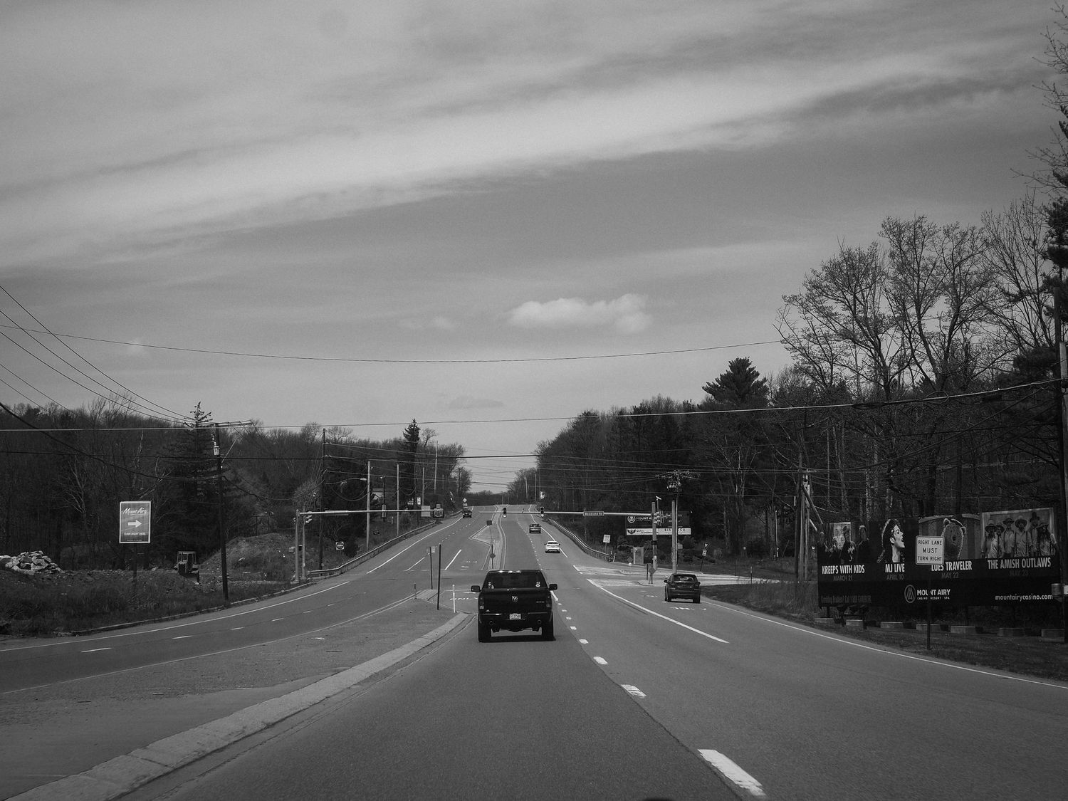 A black and white photograph shows a wide highway with power lines and trees lining both sides on a cloudy day.