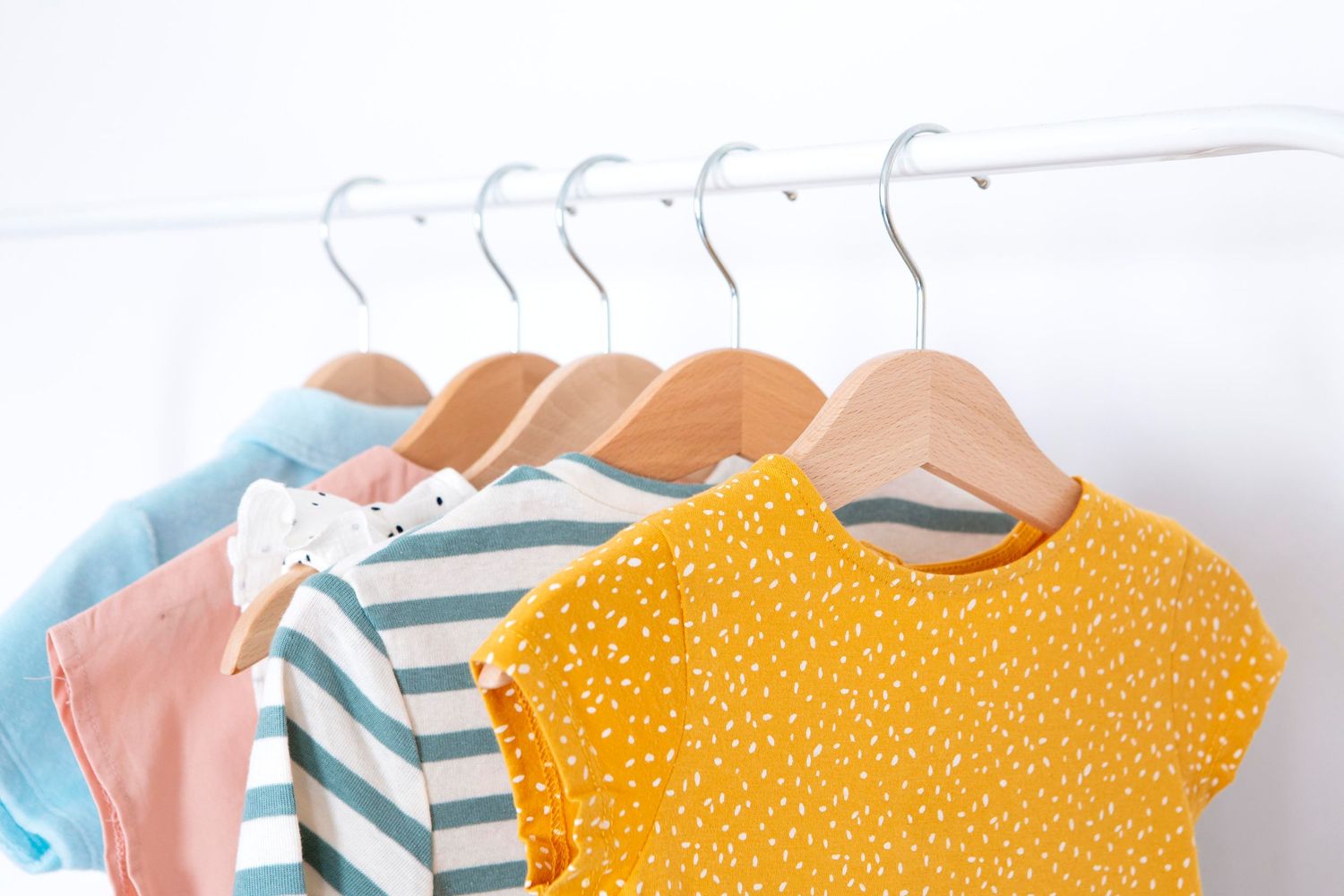 Row of colorful children's t-shirts hanging on white hangers featuring stripes, polka dots, and solid patterns.