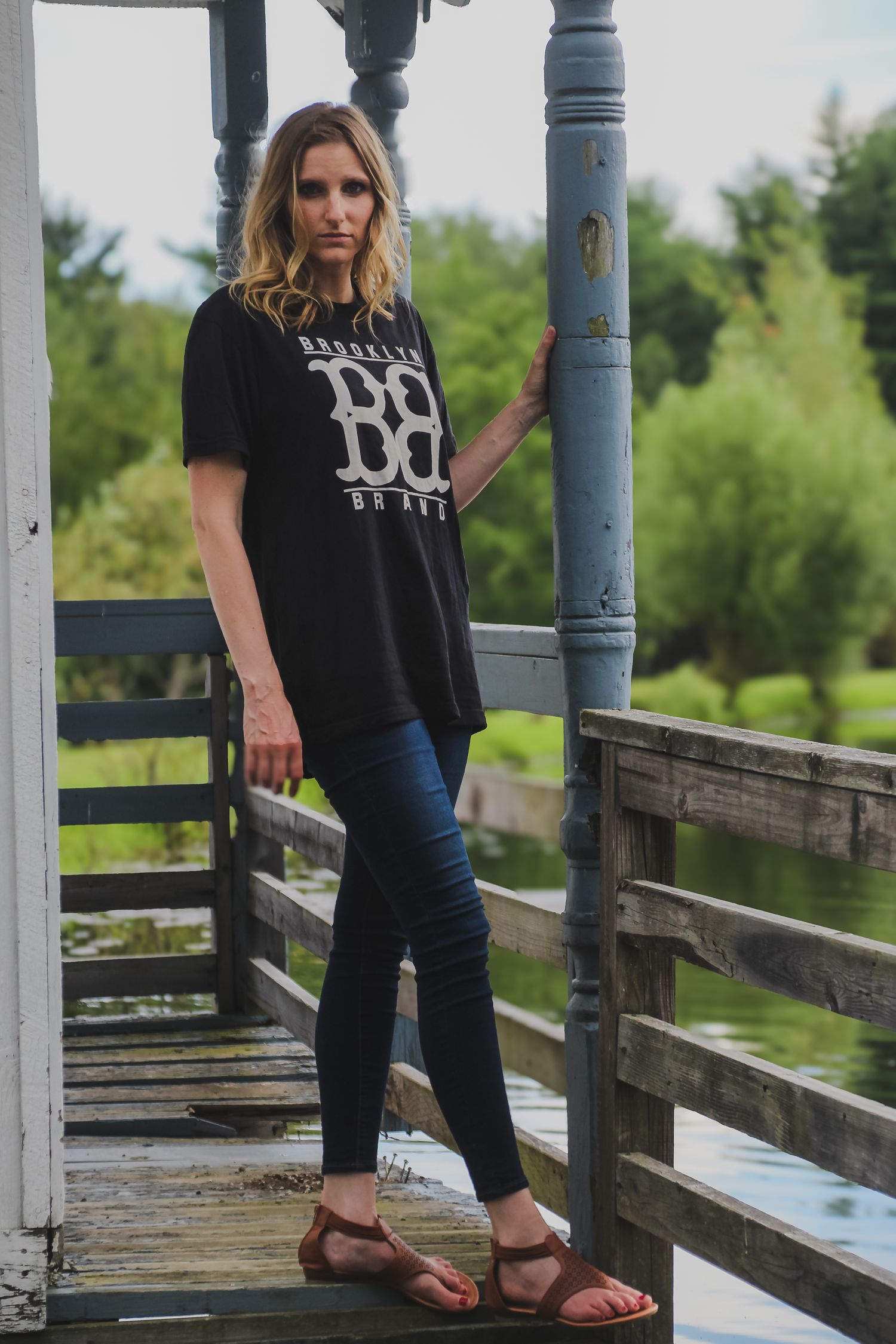 Woman in black graphic t-shirt and jeans stands on wooden boardwalk in nature setting.
