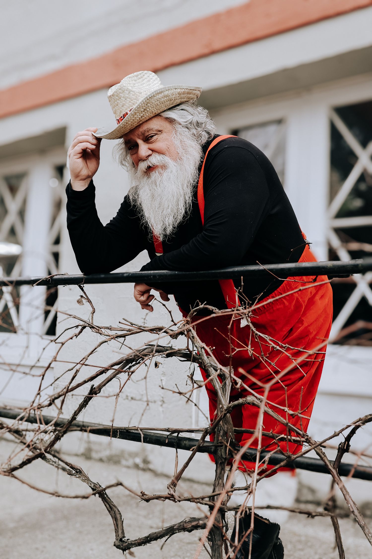 A person in a red coat and hat leans down to examine bare winter branches against a white building exterior.