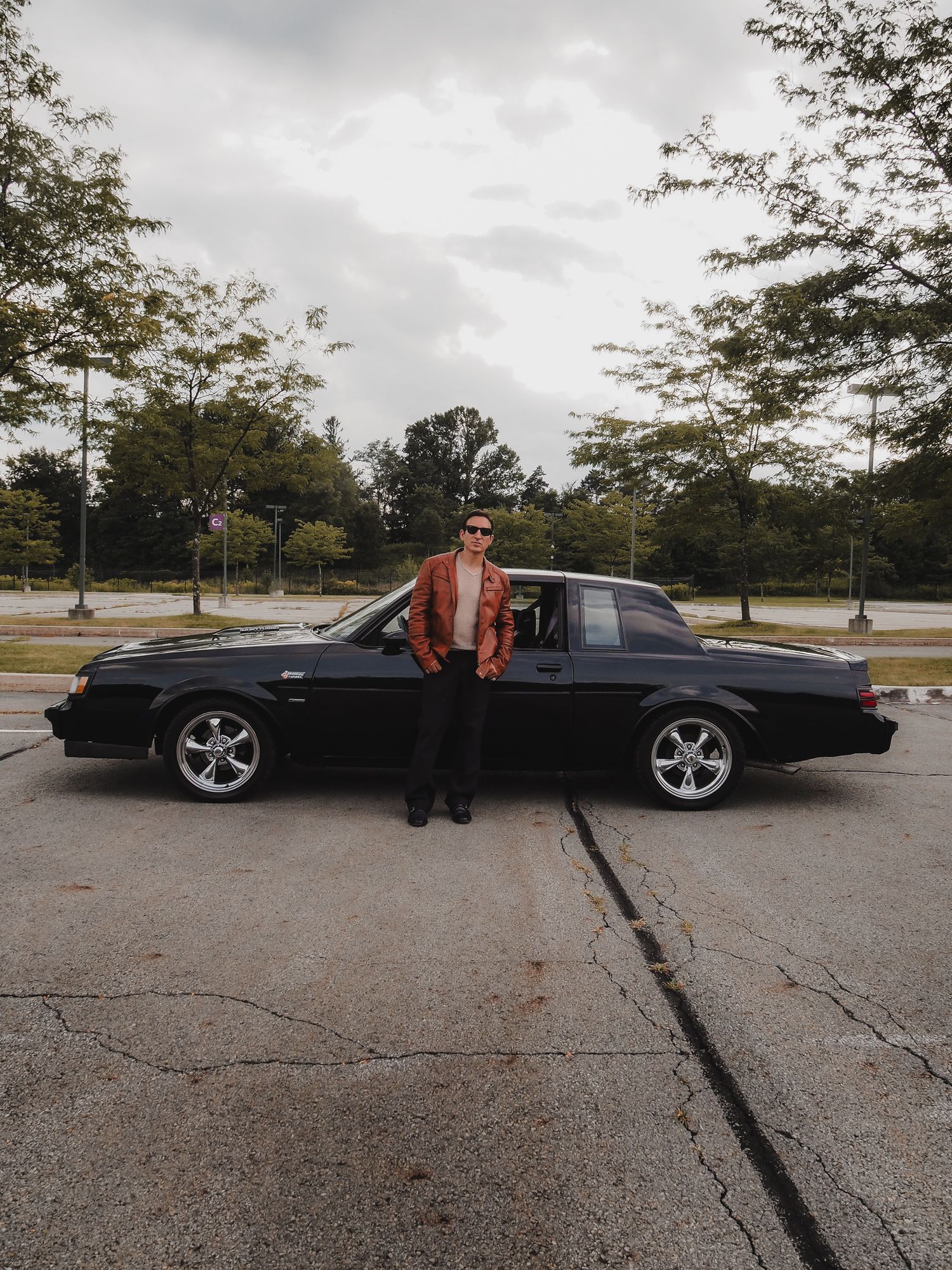 A black classic car is parked in an empty lot on an overcast day with someone standing beside it.