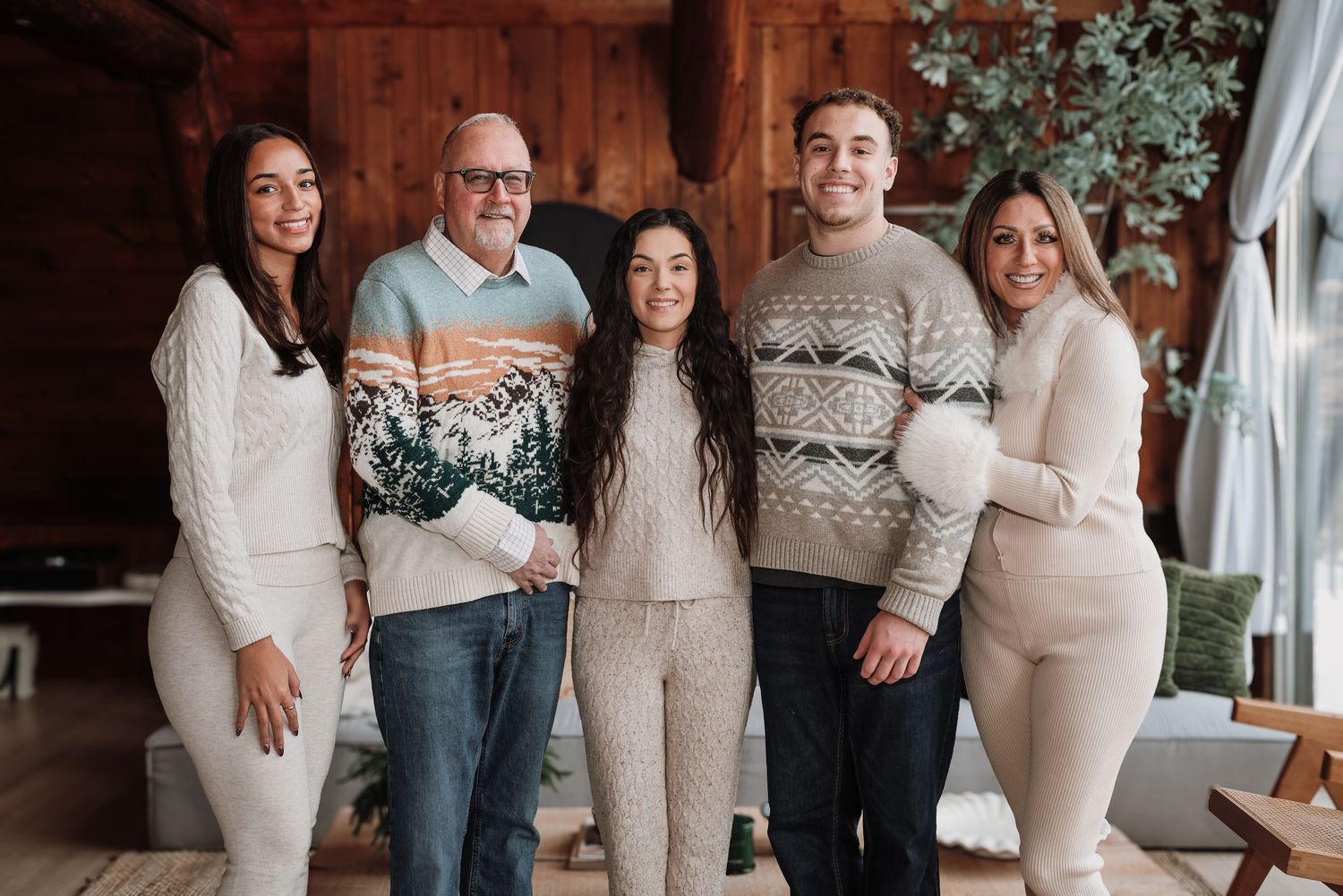A family wearing coordinated cream and beige winter sweaters poses together in a cozy wooden interior setting.