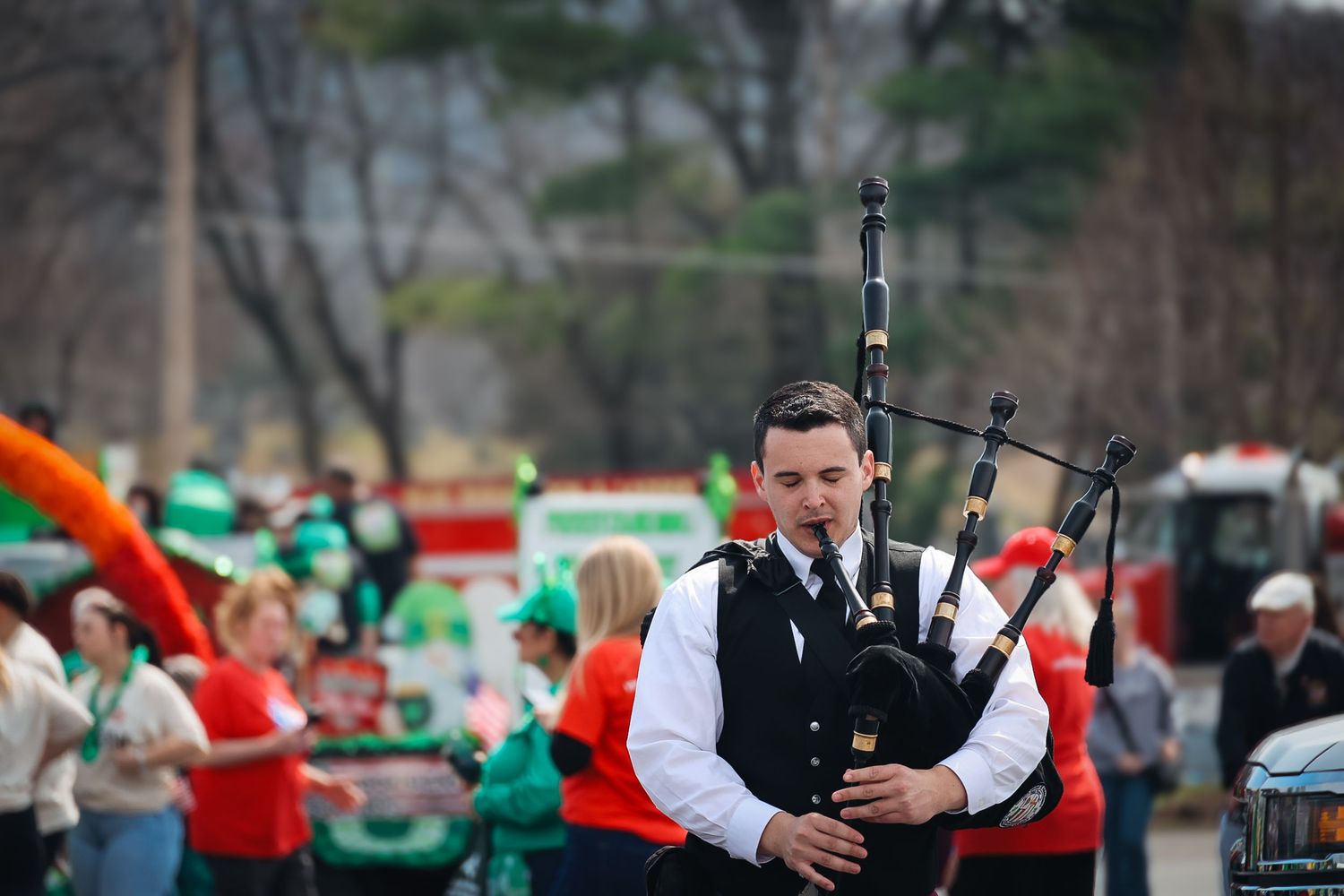 Bagpiper in black uniform performs at a St. Patrick's Day parade with crowd in green attire in background.