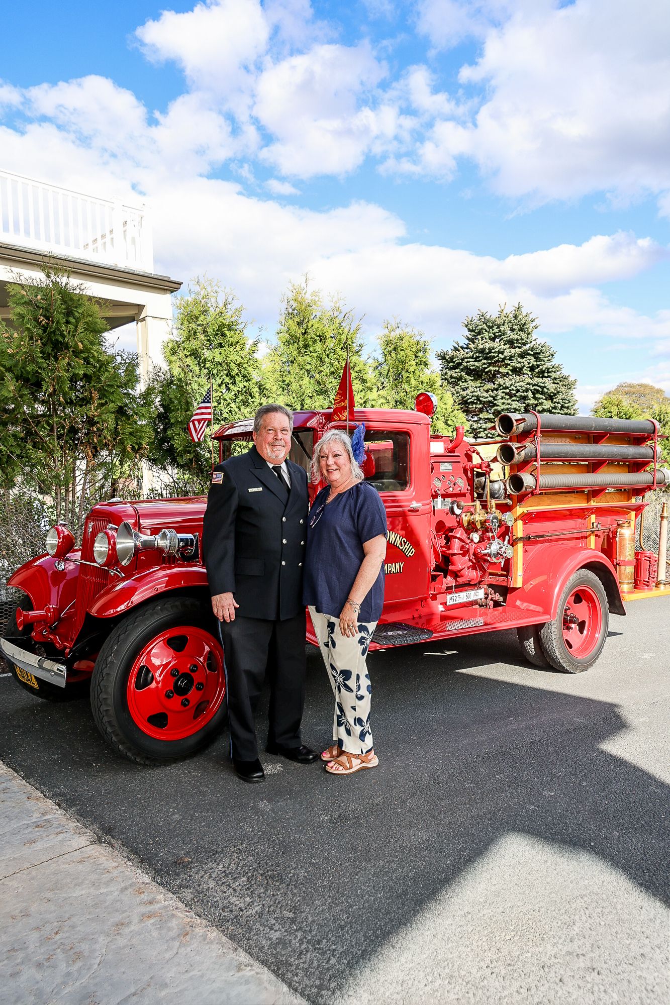 A man in uniform and a woman pose smiling in front of a vintage red fire truck outdoors on a sunny day.