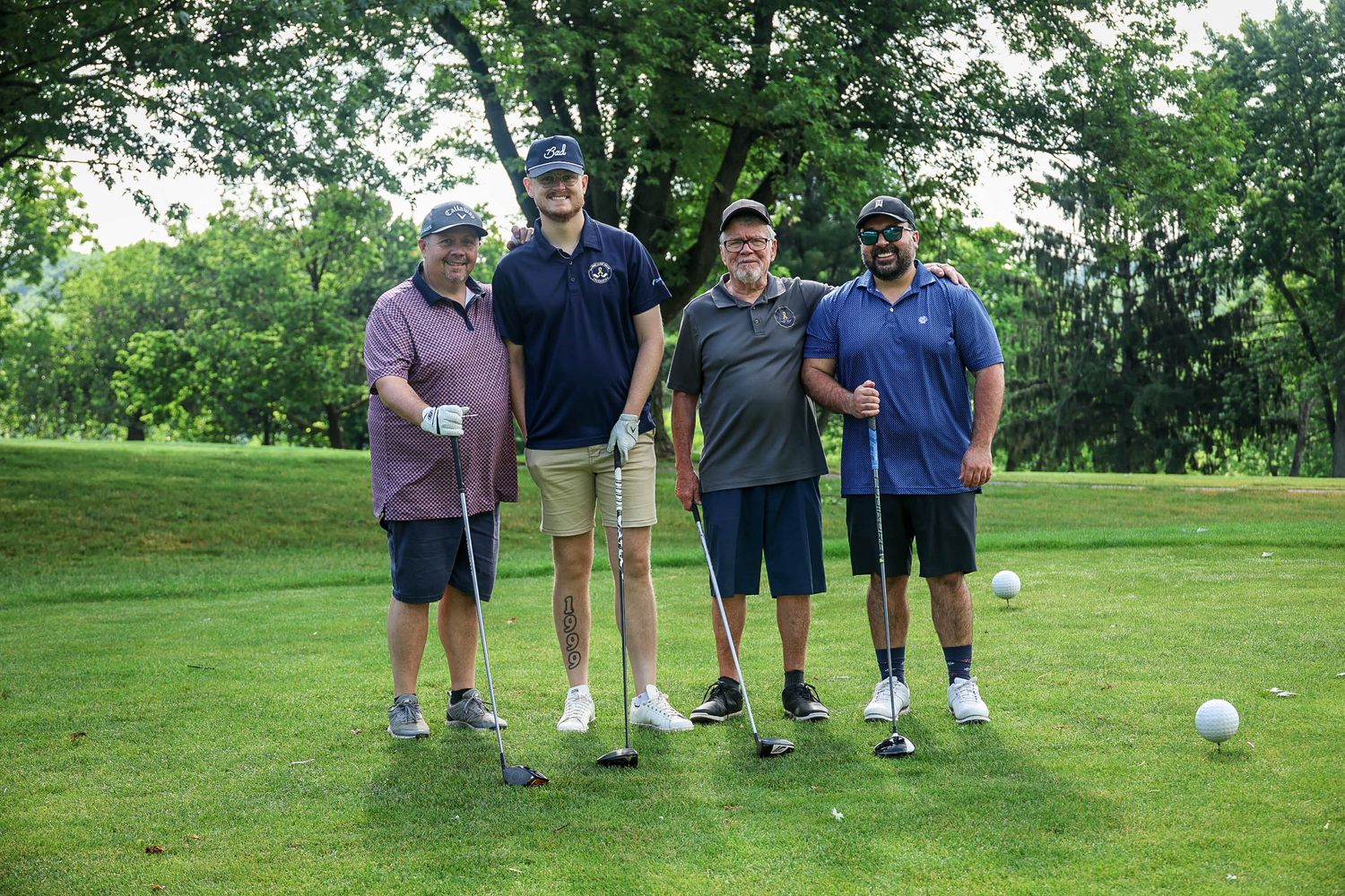 Four men posing with golf clubs on a lush green golf course on a sunny day.