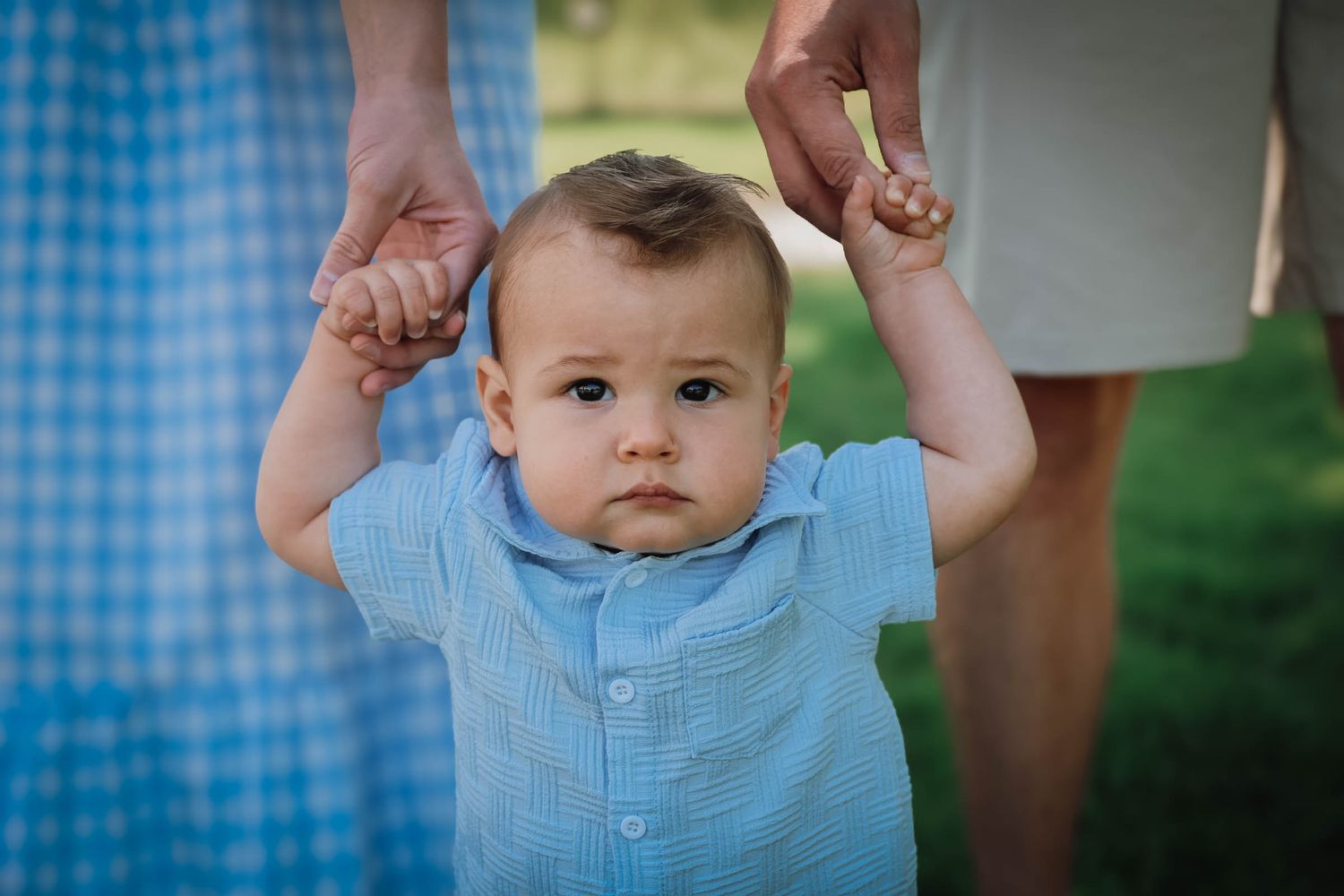 A baby boy in a blue shirt takes his first steps while being supported by two adults holding his hands outdoors.