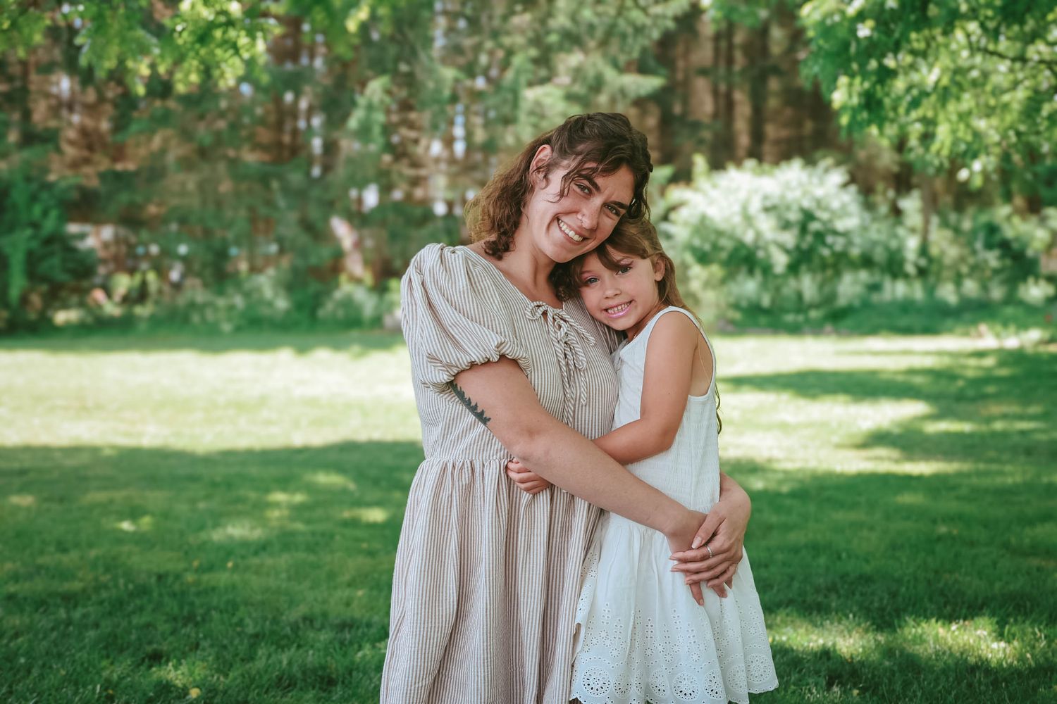 A smiling mother hugs her young daughter in a lush green garden, both wearing light beige dresses.
