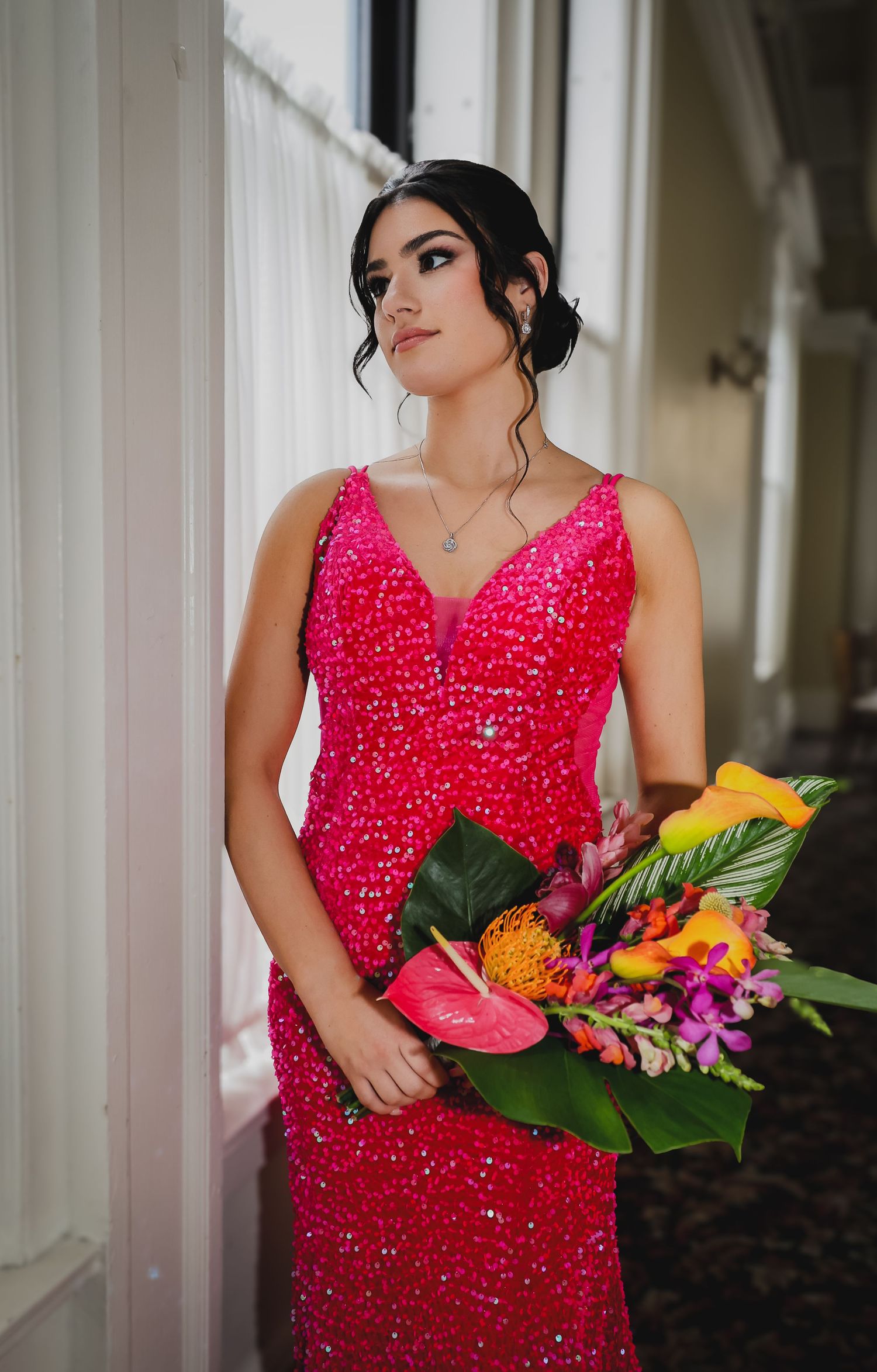Young woman in a pink sequin gown holds a tropical bouquet, posing elegantly by a window in soft natural light.