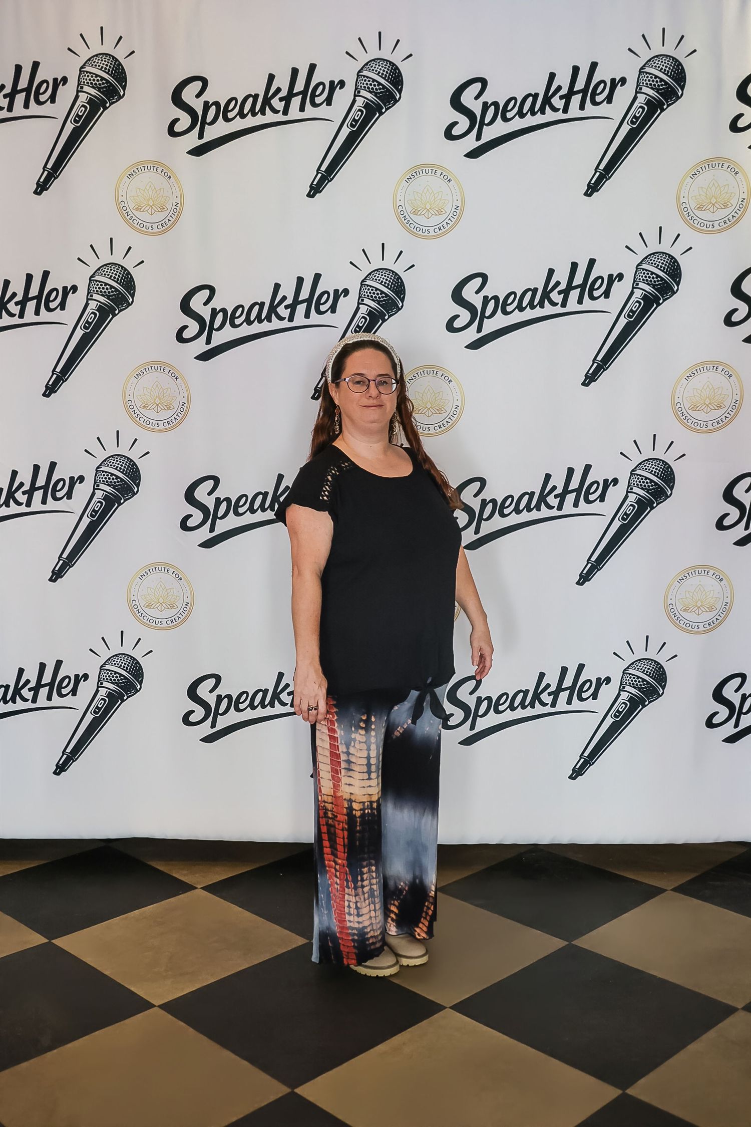 Woman in black top and tie-dye pants posing at SpeakHer event step-and-repeat backdrop.