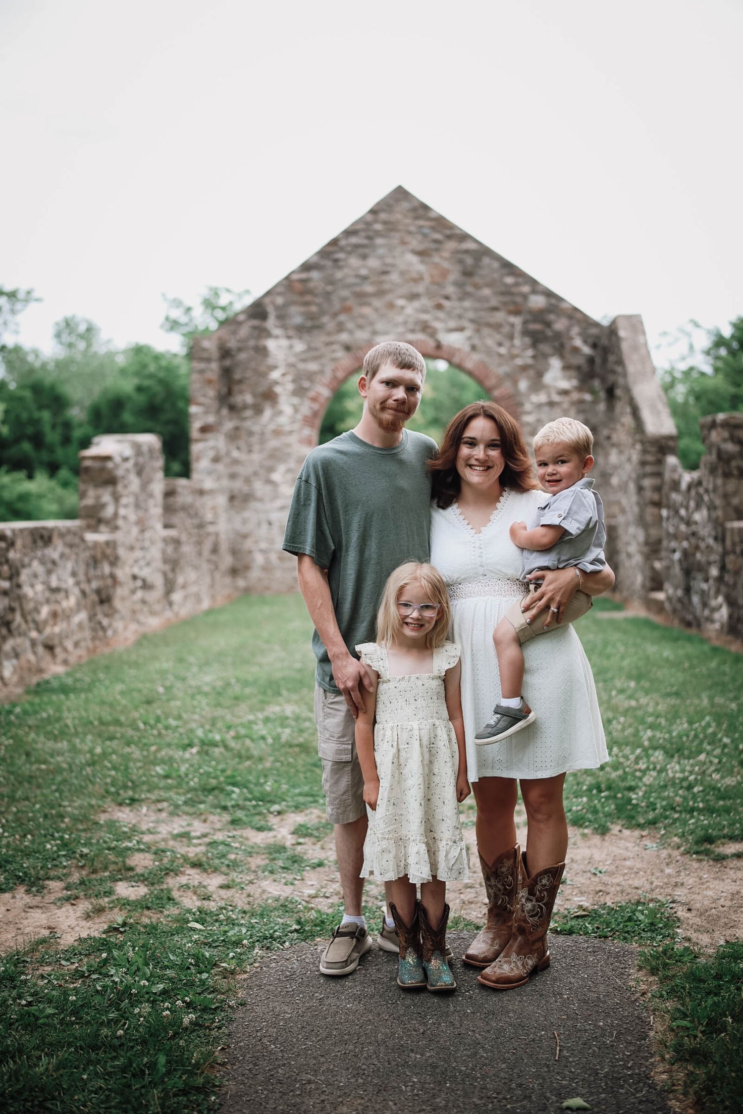 Family of four posing in front of stone ruins, parents with young daughter and baby, outdoors on green pathway.