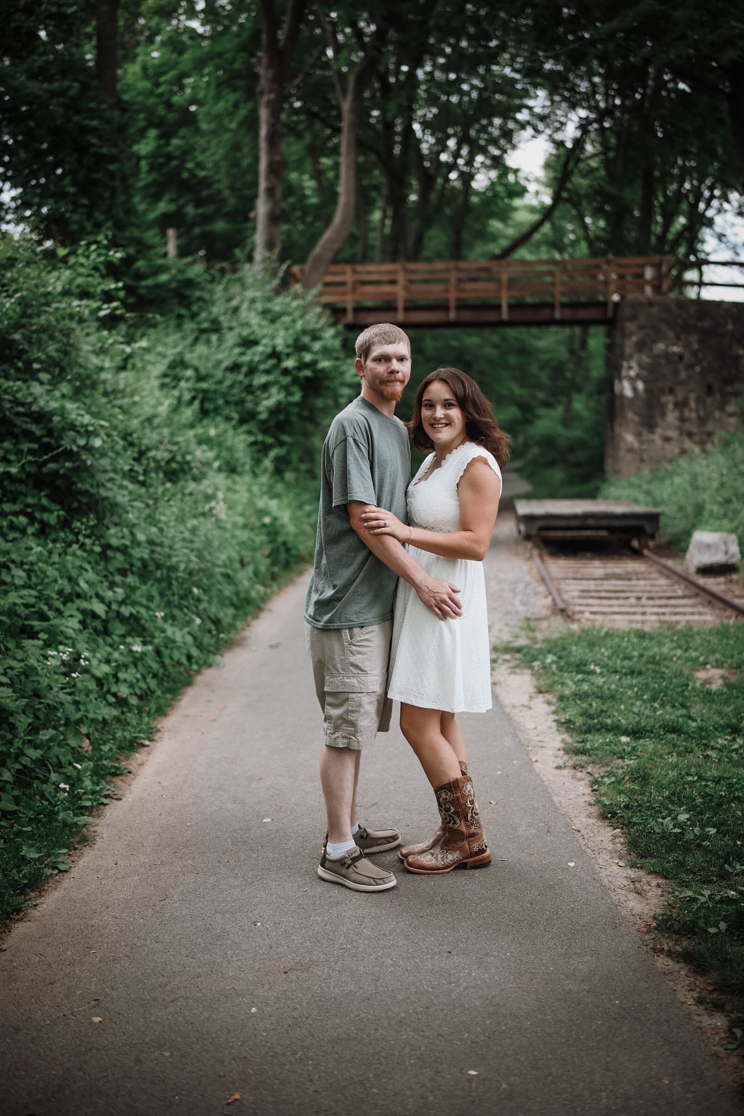 Couple embracing on a scenic park pathway surrounded by lush green trees and an old bridge in the background.