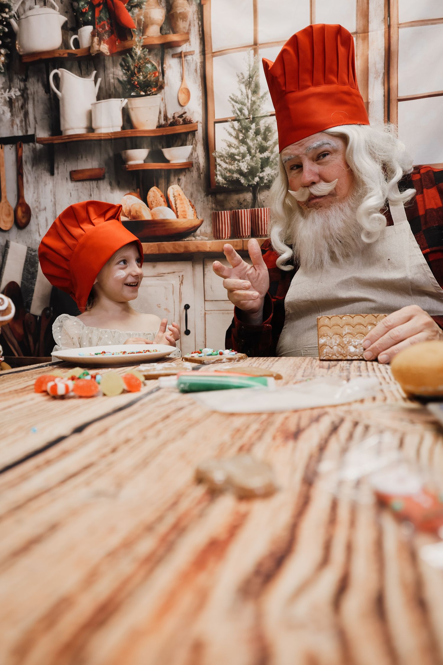 Santa Claus and child in red chef hats decorating Christmas gingerbread cookies together in a festive kitchen.