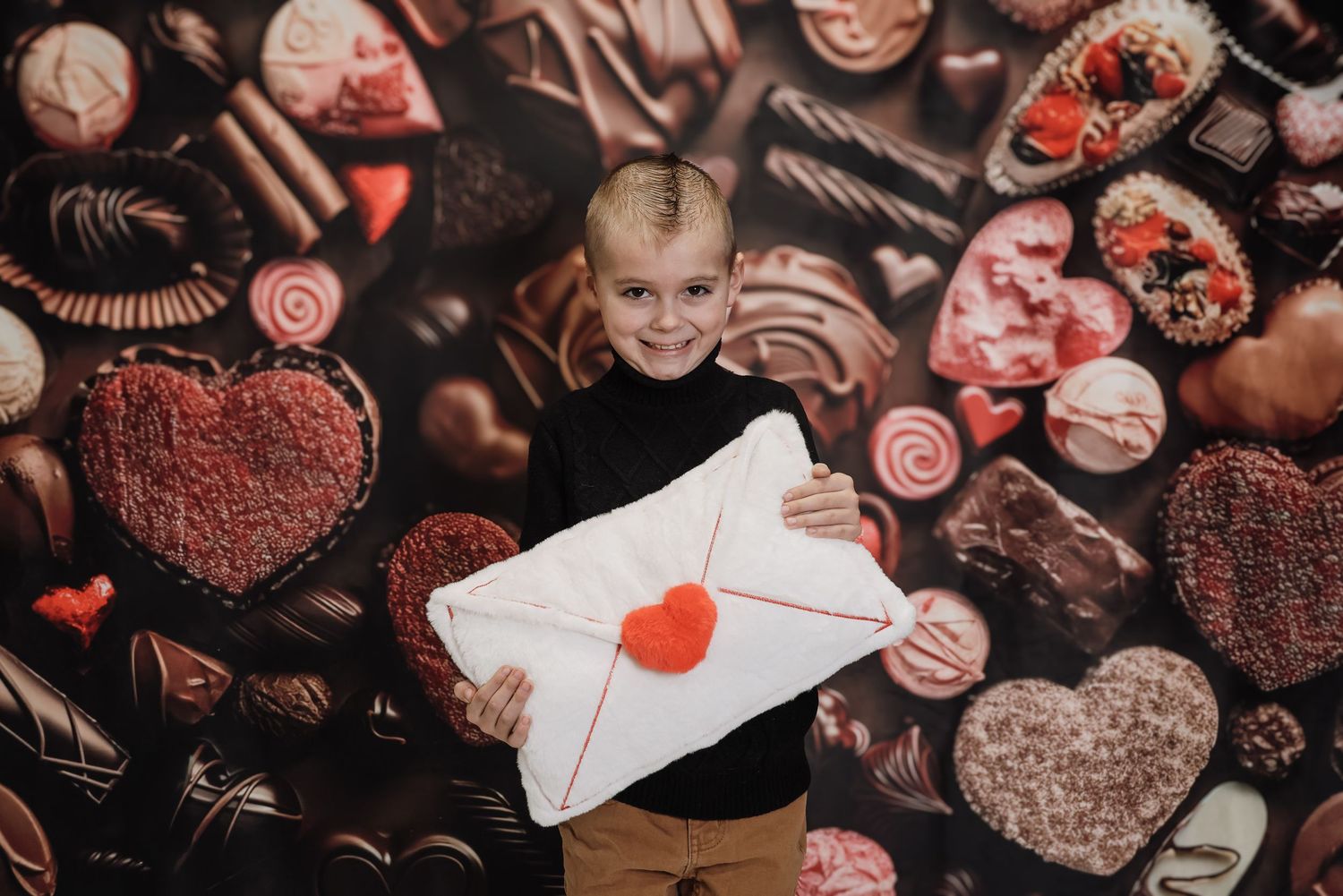 A smiling blonde child holds a white envelope with a red heart seal against a chocolate candy Valentine backdrop.