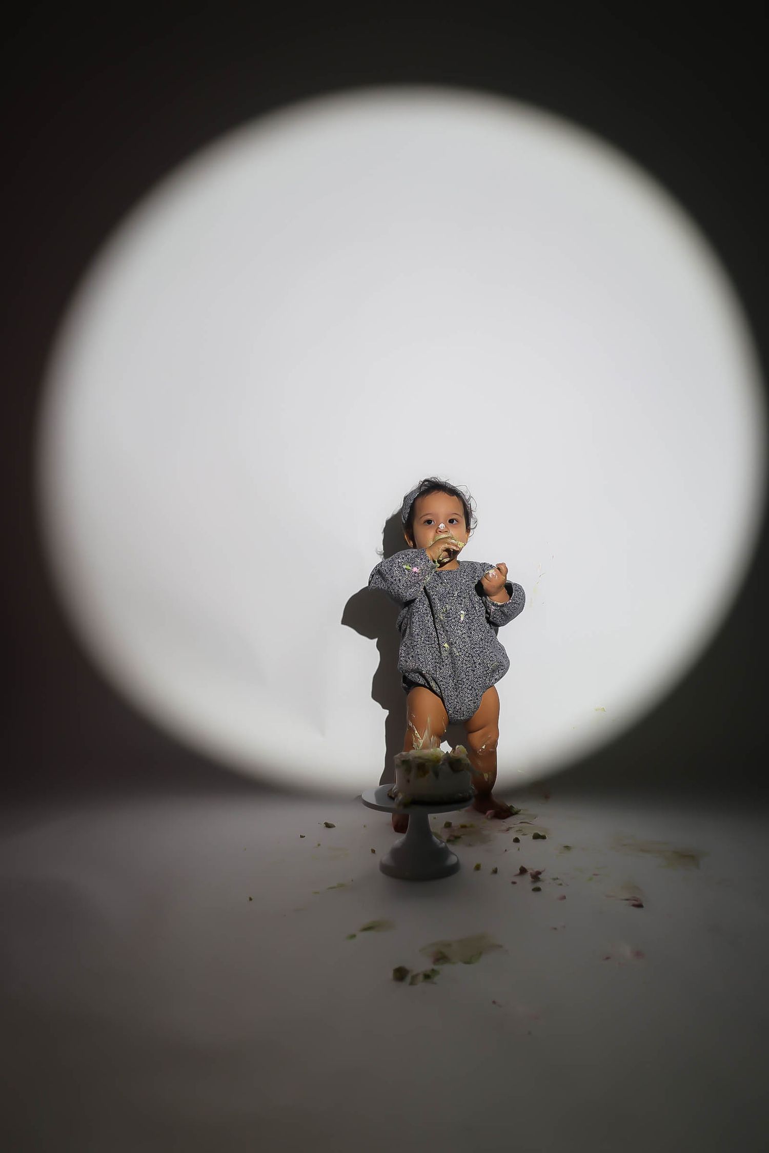 Baby stepping on smashed birthday cake in gray romper under dramatic circular spotlight.
