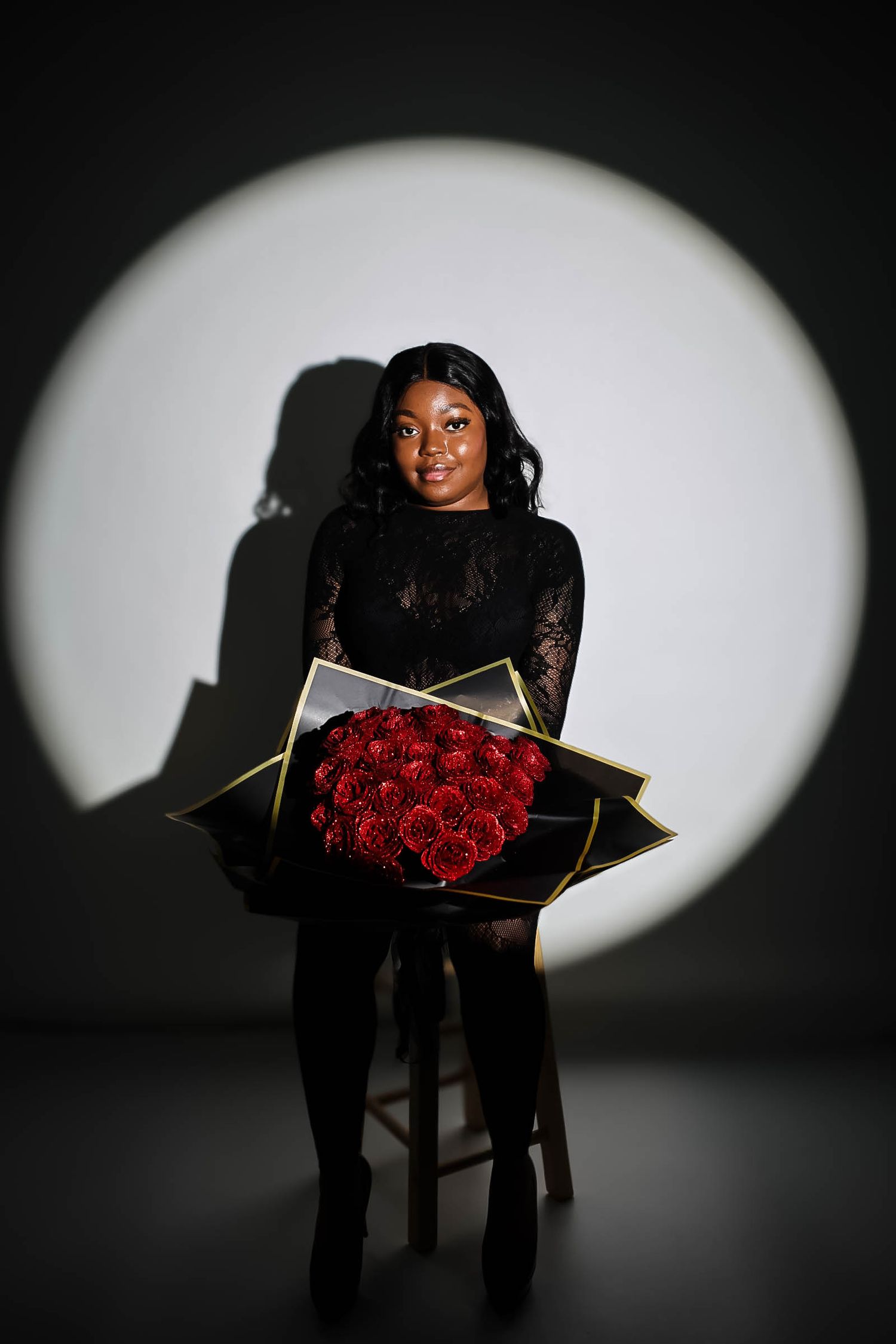 Young woman in black lace dress holding large red rose bouquet, posed against dramatic spotlight background.