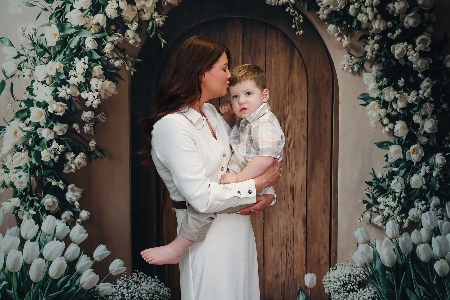 A woman in a white dress holds a young toddler boy amid lush white floral arrangements by a wooden door.