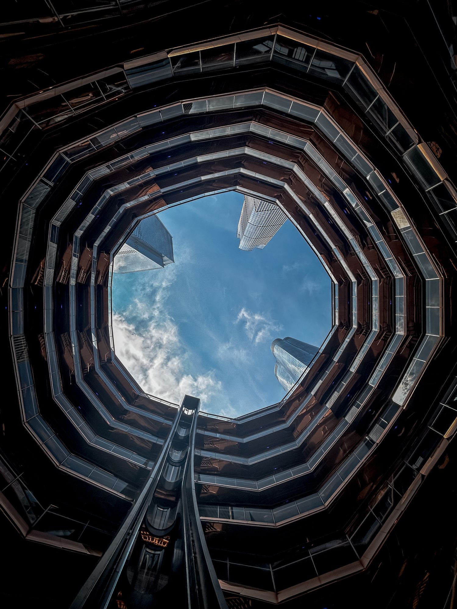 Looking up through a circular architectural opening revealing blue sky, skyscrapers, and a central escalator shaft.