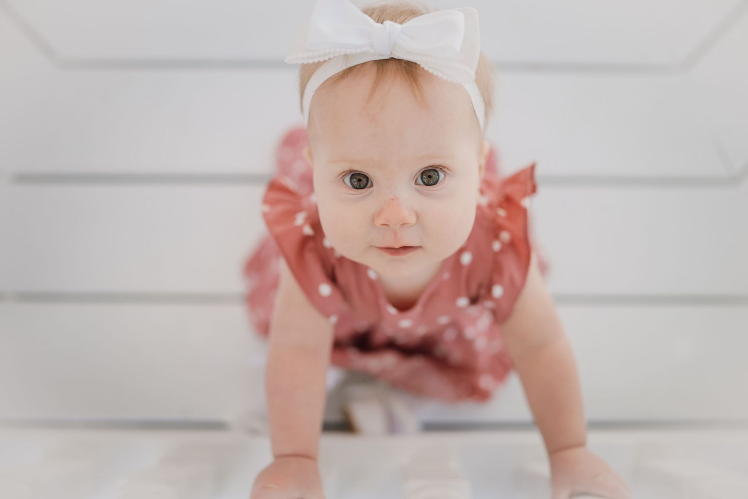 Adorable baby girl in pink polka dot dress and white bow headband crawling on white wooden floor.