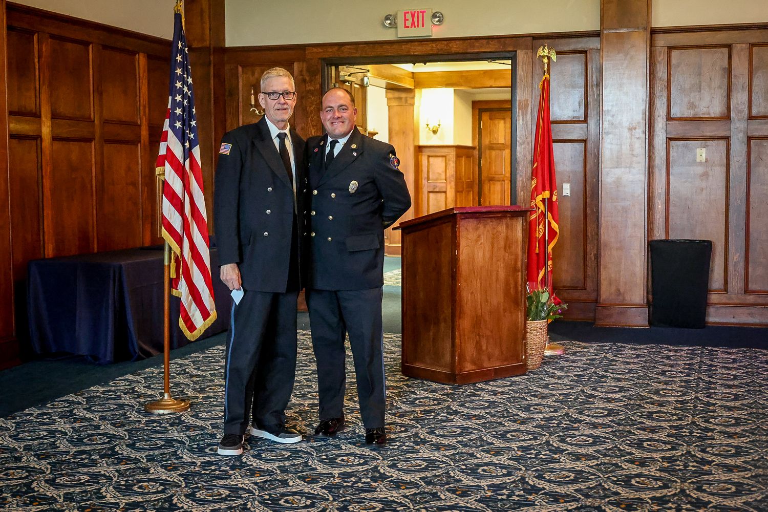 Two men in formal uniforms pose together near an American flag and podium at an official ceremony.