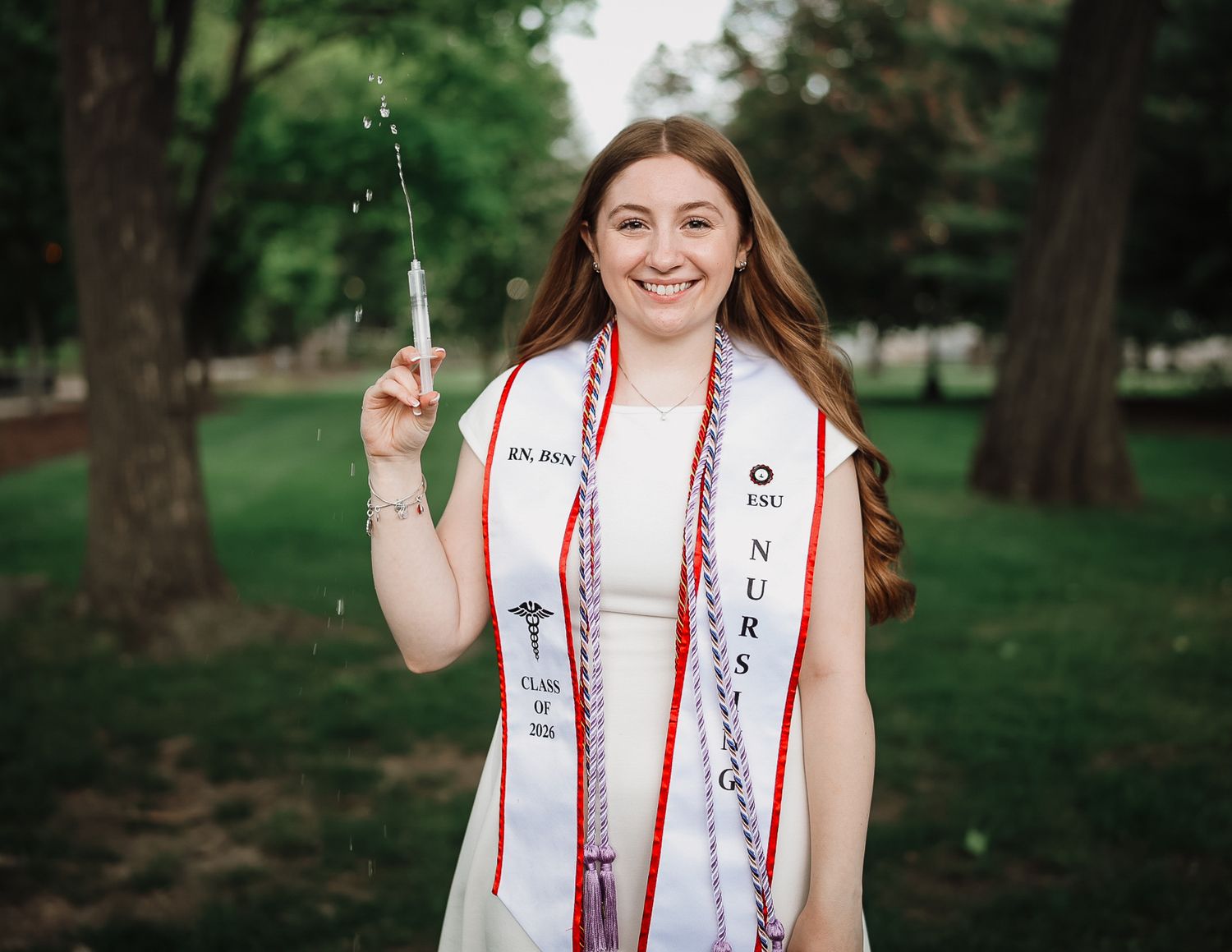 Smiling female nursing graduate holds syringe prop outdoors wearing white dress and honor stoles.
