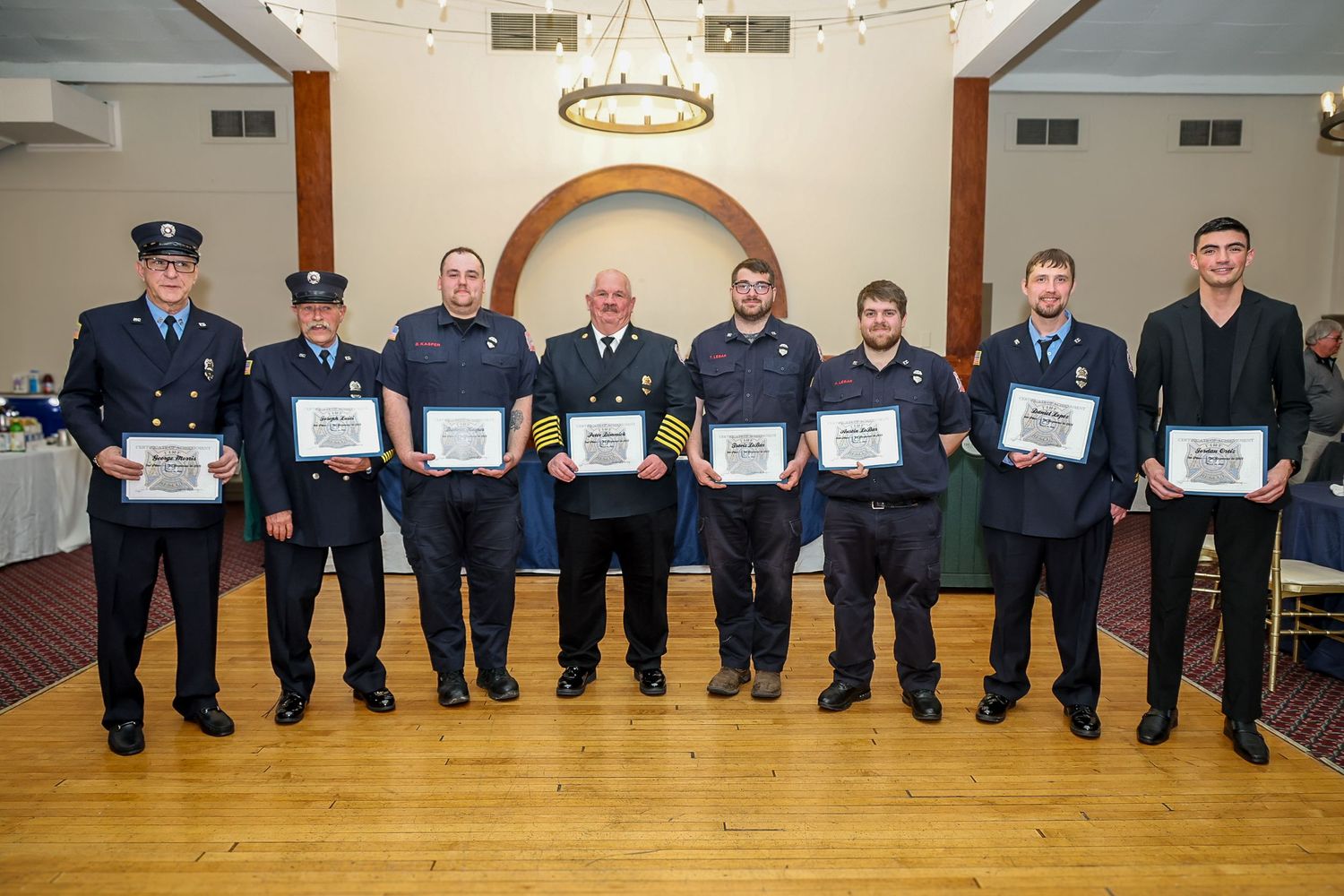 Seven firefighters in uniform proudly holding framed awards at a formal recognition ceremony indoors.