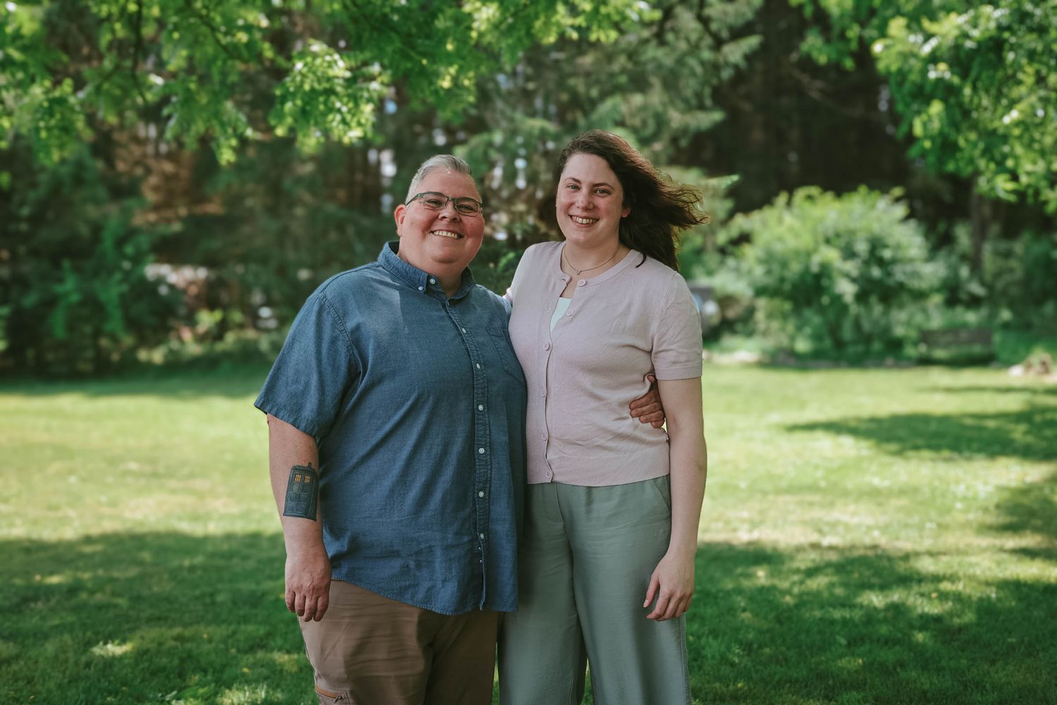 A smiling man and woman stand together outdoors on a sunny day in a lush green garden.