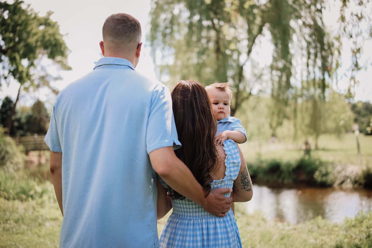 A family of three stands outdoors near a pond, parents in blue outfits holding a baby, surrounded by lush green trees.