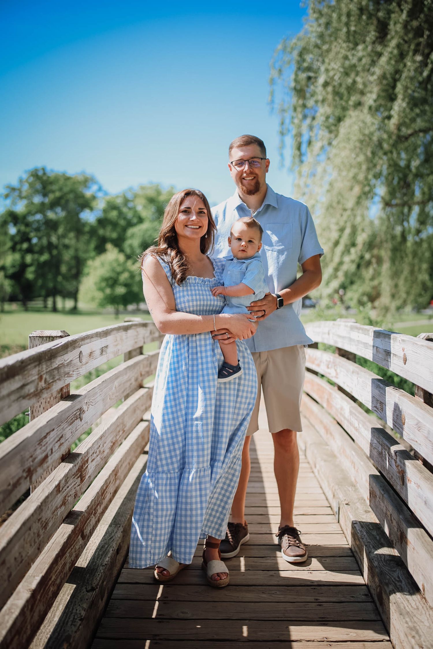 Young family with baby posing on wooden bridge in sunny park, wearing blue outfits, smiling outdoors.