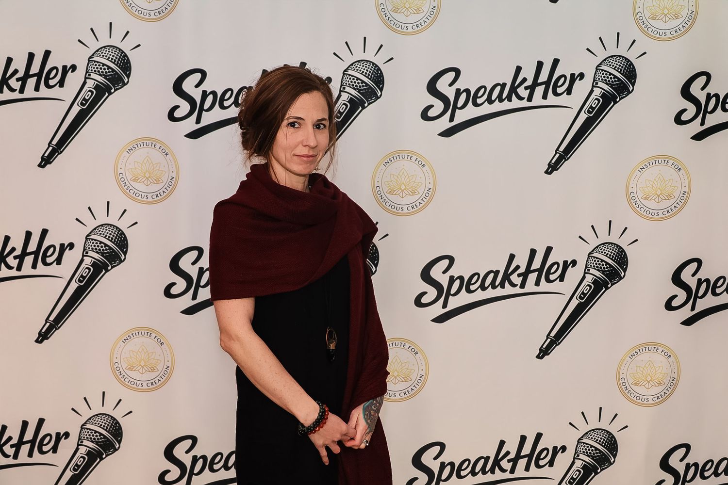 Woman in black dress and burgundy scarf poses at SpeakHer event backdrop with microphone logos.