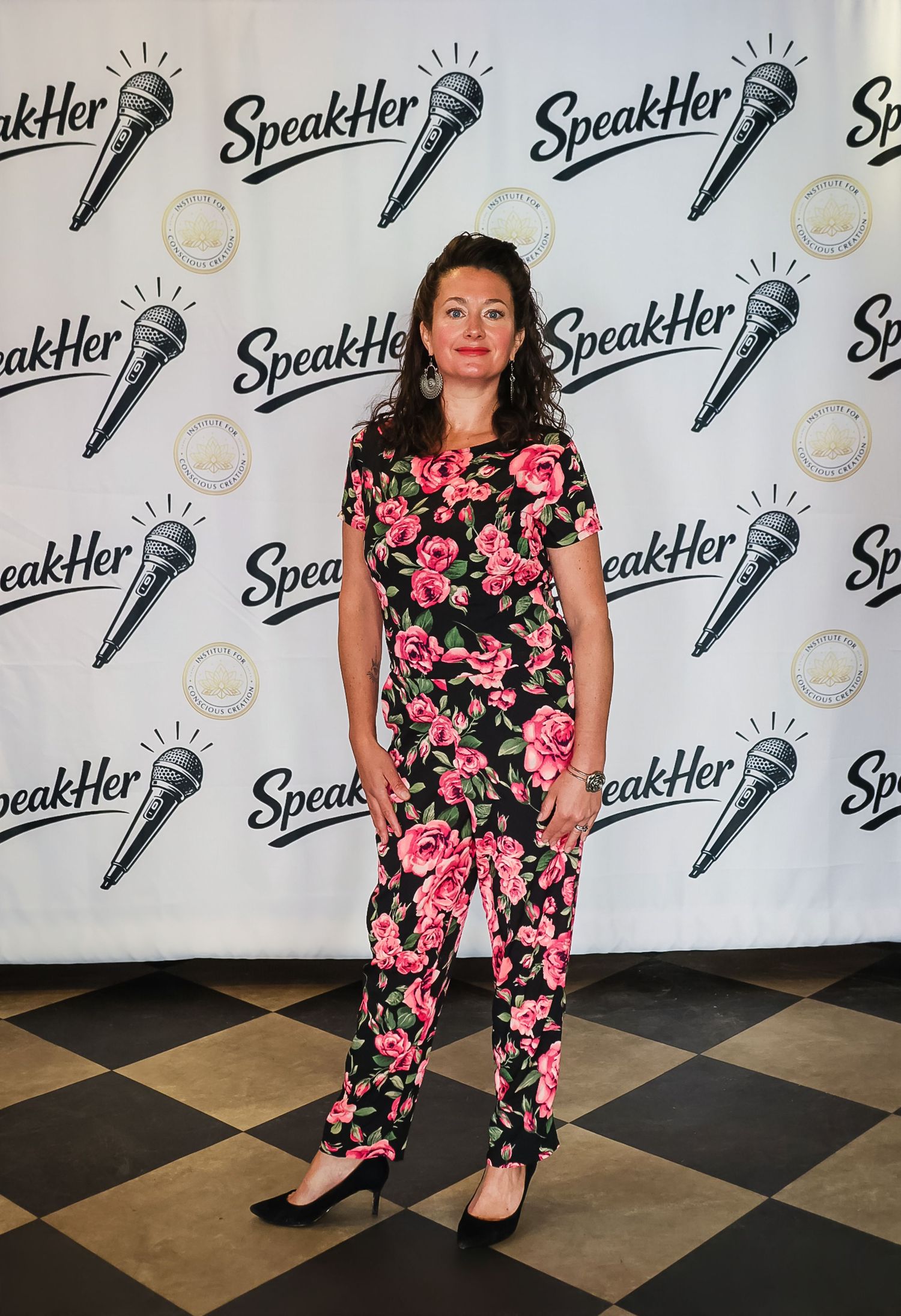 Woman in floral jumpsuit posing at SpeakHer event backdrop with microphone logos.
