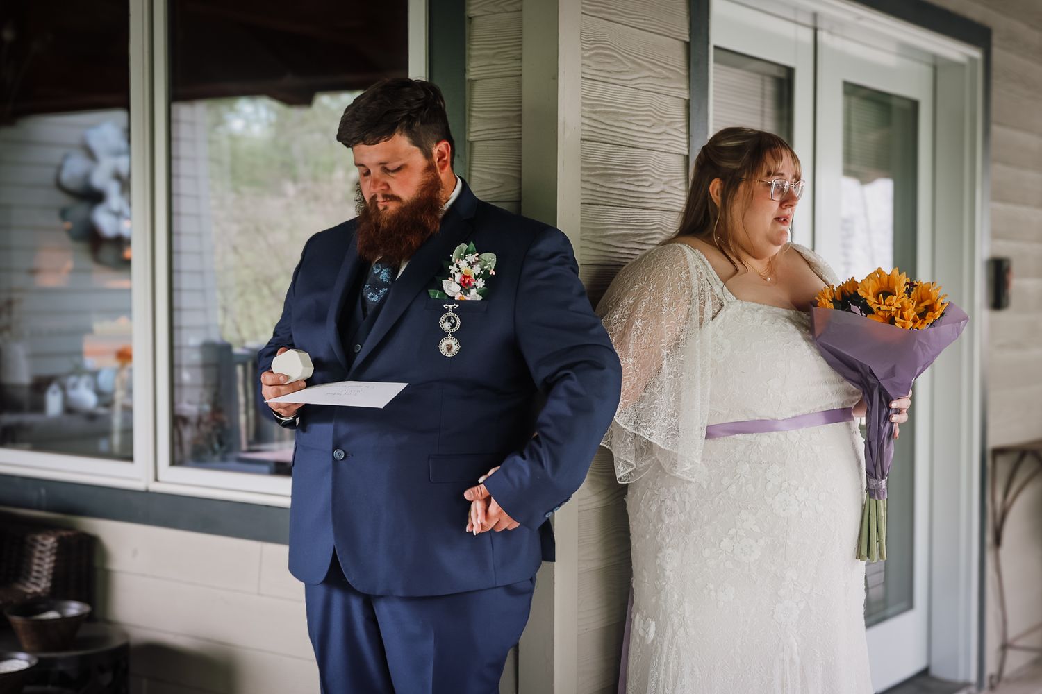 Groom in navy suit reads vows while bride in white gown holds sunflowers during outdoor first look wedding moment.