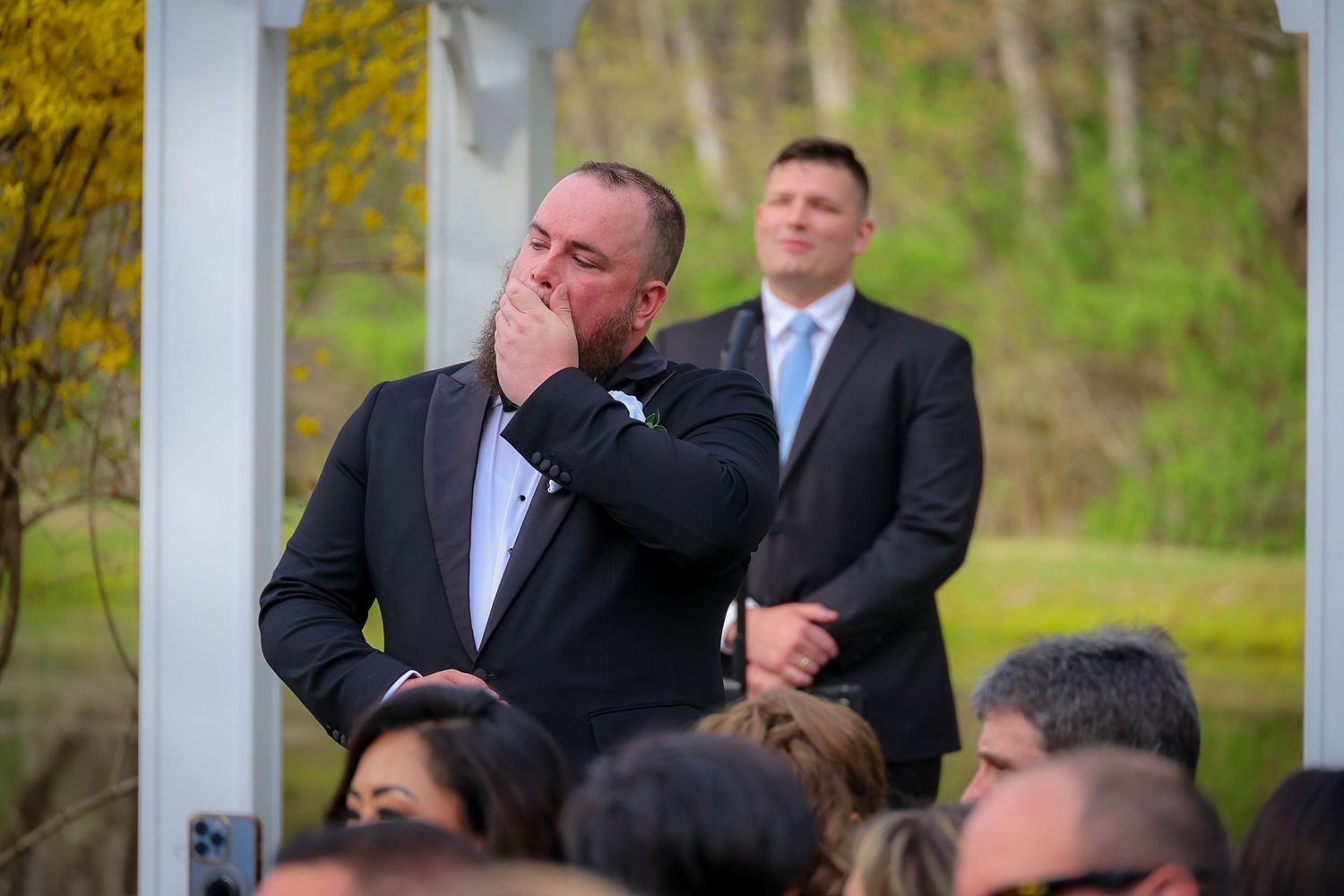Emotional groom covers mouth in tears during outdoor wedding ceremony with guests and arbor in background.