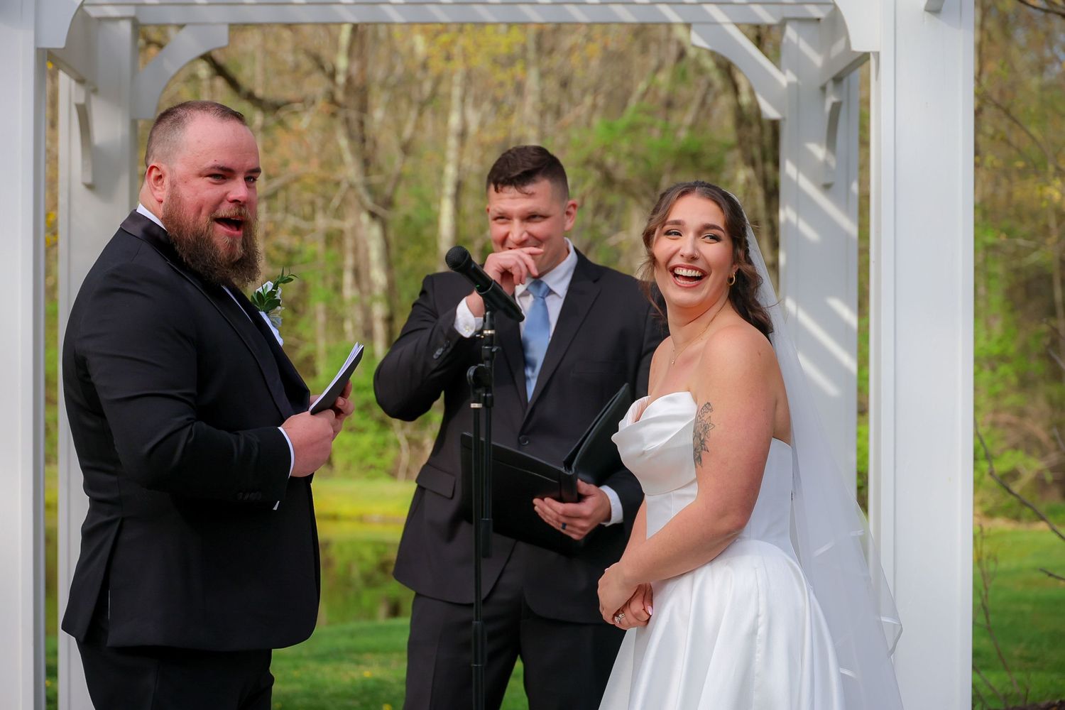A laughing bride and groom exchange vows outdoors under a white arbor with an officiant during a spring wedding ceremony.