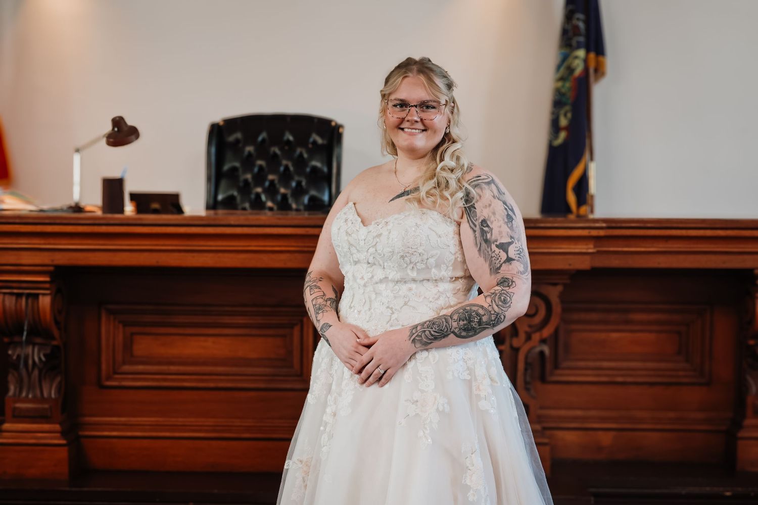 Tattooed bride in lace A-line wedding dress smiling in a wood-paneled courthouse with a New Hampshire flag.