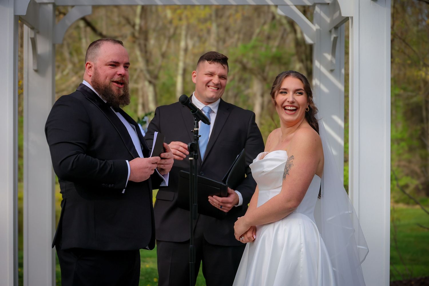 Bride and groom exchange vows with officiant under white pergola at outdoor wedding ceremony.