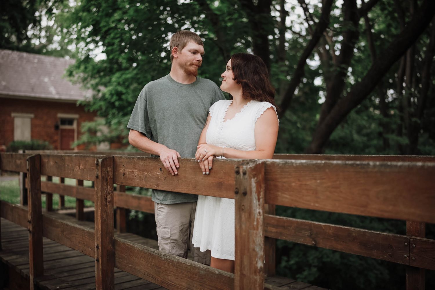 Couple standing on a rustic wooden bridge surrounded by lush green trees, sharing a romantic moment outdoors.
