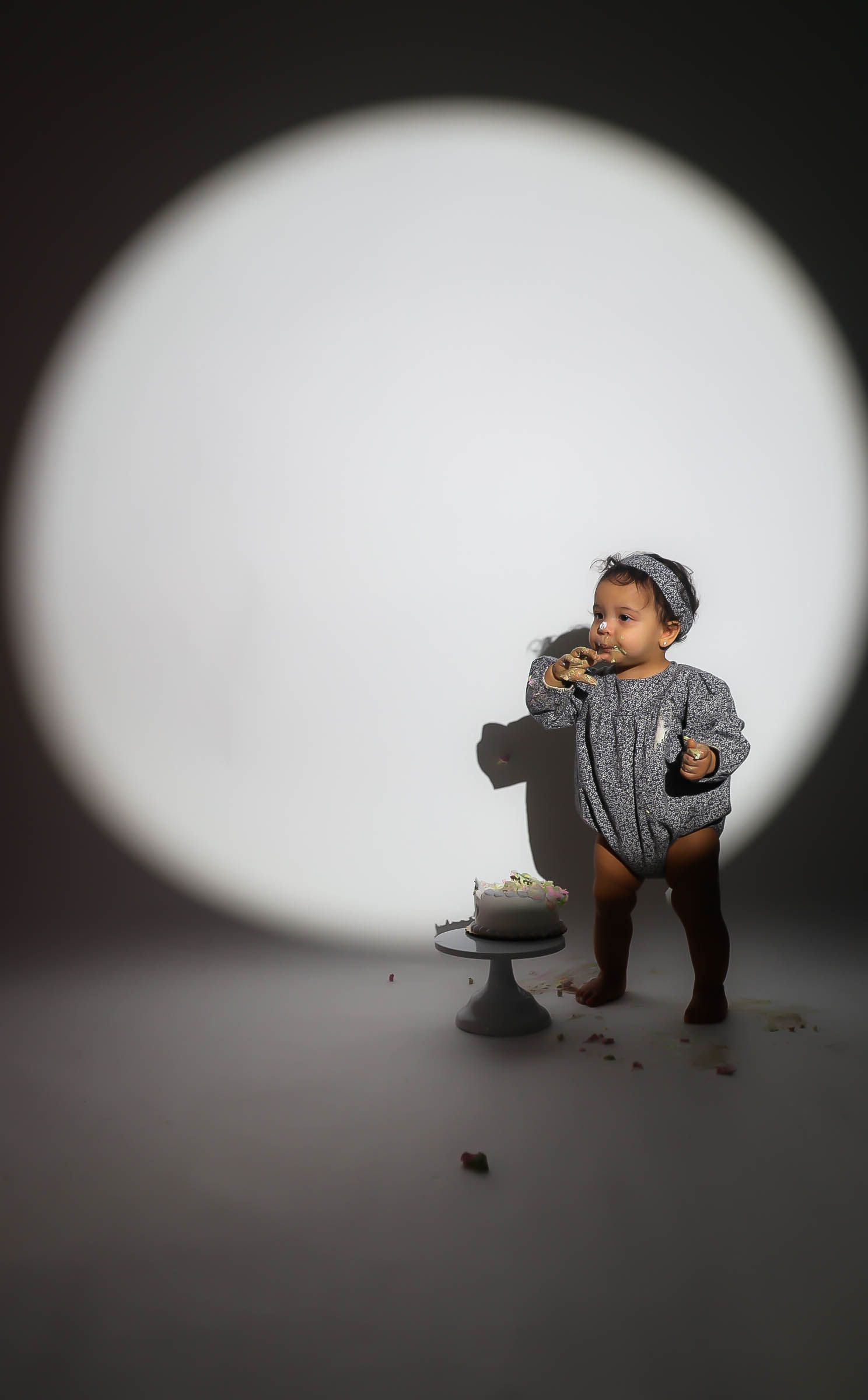 A baby girl in a floral romper eats cake beside a cake stand under a dramatic circular light.
