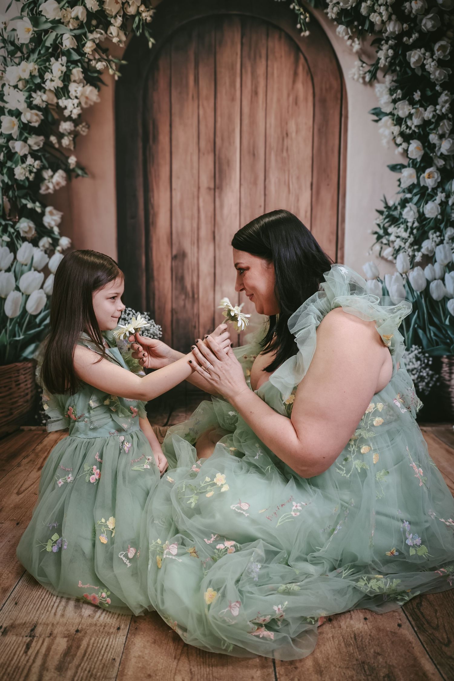 Mother and daughter in matching sage green floral tulle dresses sharing flowers in a whimsical floral studio setting.